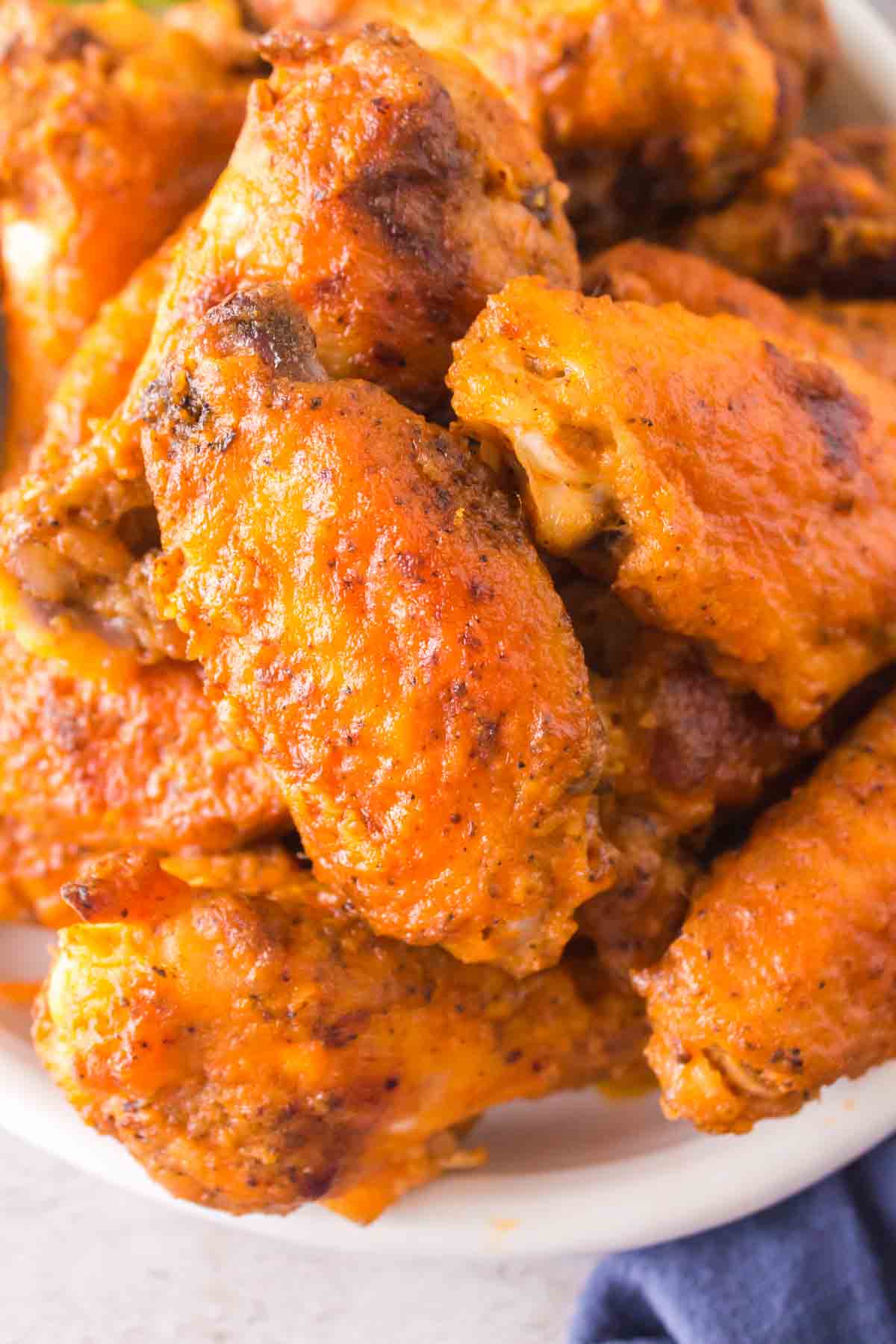 A close-up of a plate of hot wings showcases the buffalo chicken wings coated in a bright orange, spicy sauce, with a golden-brown, crispy texture. The background hints at a blue cloth, providing contrast to the dish.