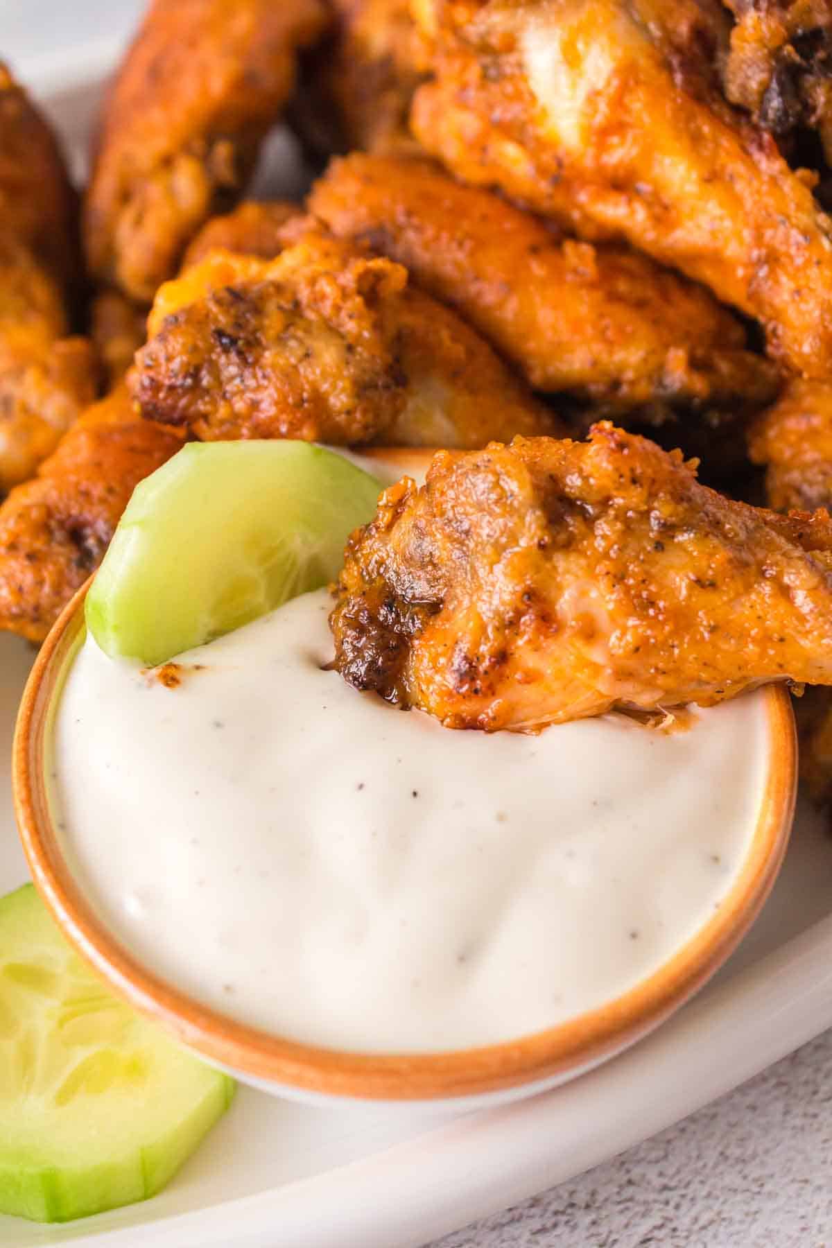 A close-up of crispy, golden-brown chicken wings placed next to a small bowl of creamy white dip with black specks. Two cucumber slices are next to the dip, all served on a white plate.