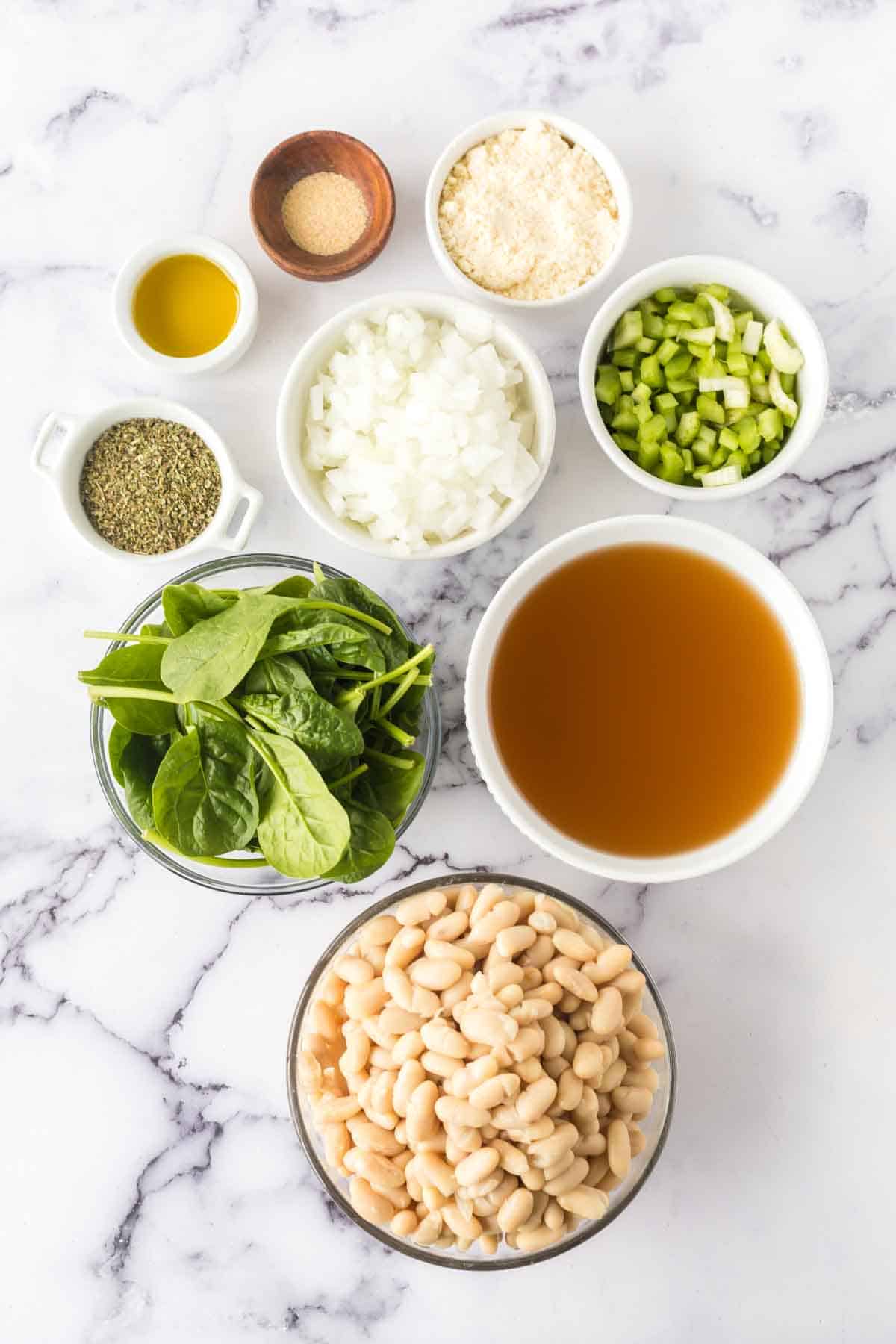 On a marble surface, the ingredients for white bean soup await: fresh spinach, white beans, chopped celery and onions, with vegetable broth ready to simmer. Grated cheese, olive oil, dried herbs, and granulated garlic sit poised in separate bowls to complete the savory ensemble.