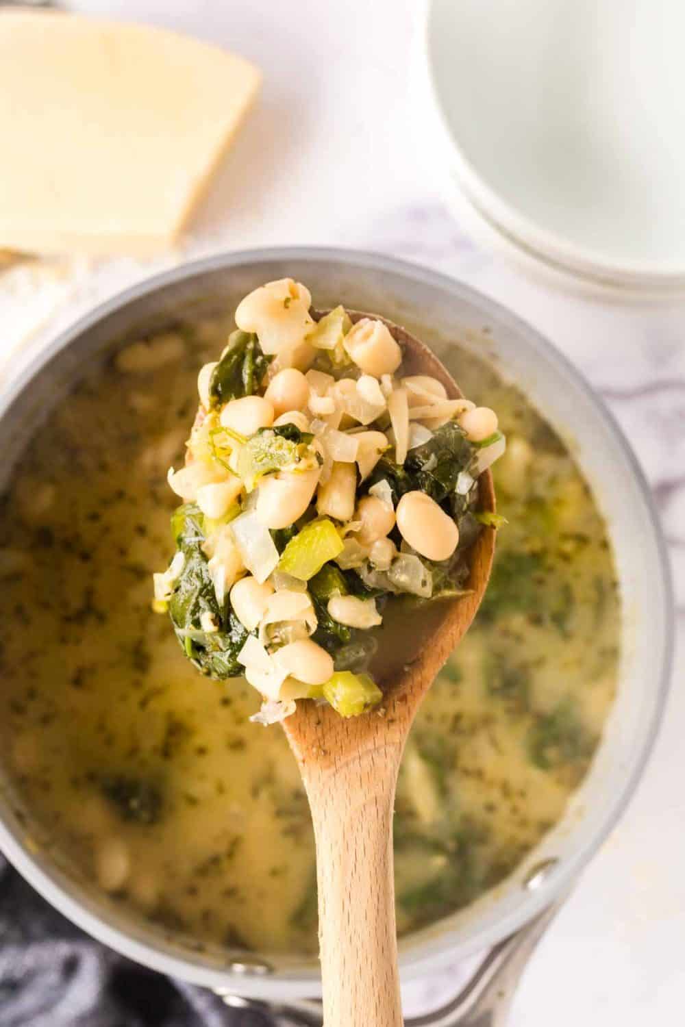 A wooden spoon holds a portion of white bean soup above a pot. The soup features hearty white beans, fresh greens, and chopped vegetables in a clear broth. In the background, a block of cheese and empty bowls await their turn to join the meal.