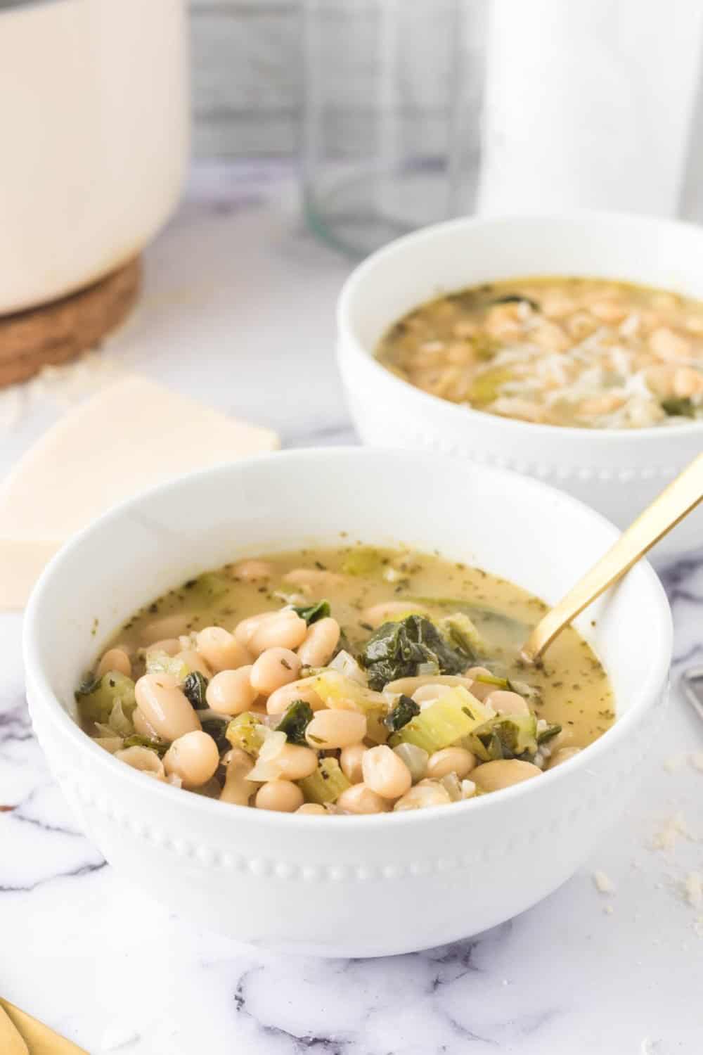 Two white bowls filled with white bean soup featuring greens and herbs. The soup is garnished with a sprinkle of cheese. A gold spoon rests in the foreground bowl. The setting includes a marble surface and a blurred background, highlighting the comforting dish.