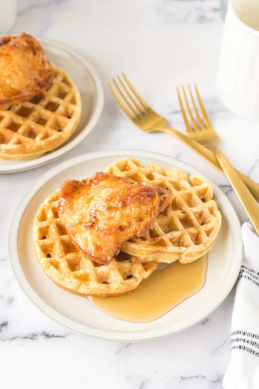 Plates of chicken and waffles, made from a beloved chicken and waffles recipe, are topped with syrup on a marble countertop. Two forks rest beside the dish, and a white and black striped napkin is nearby. Another plate with the same delicious meal is in the background.