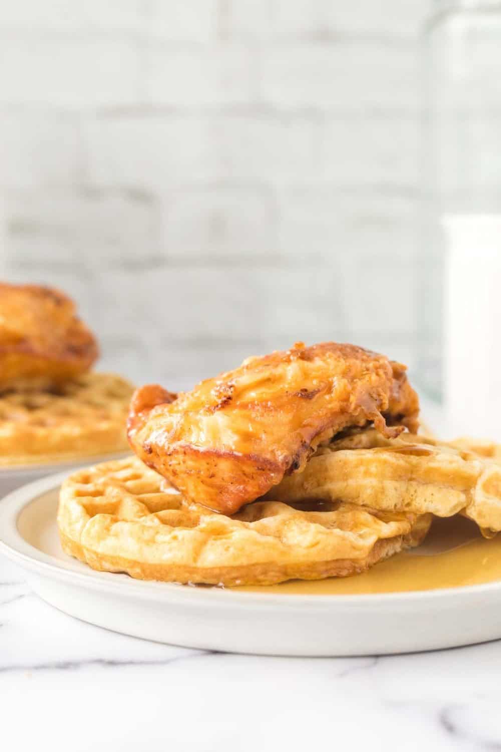 A serving of crispy fried chicken is nestled on two waffles, drizzled with syrup, atop a white plate. In the background, another chicken and waffles plate and a glass of milk complete the cozy scene against a light brick wall.