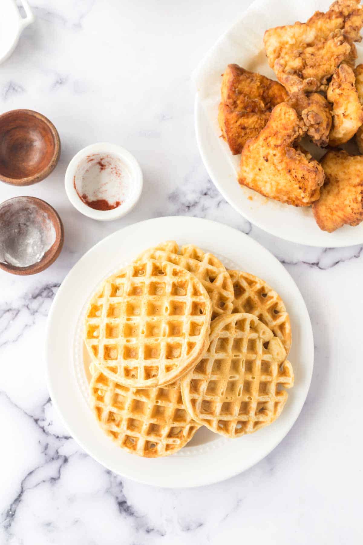 A plate of golden waffles sits on a marble surface, perfectly paired with crispy fried chicken. Nearby, small wooden bowls enhance the inviting setup for a classic chicken and waffles meal.