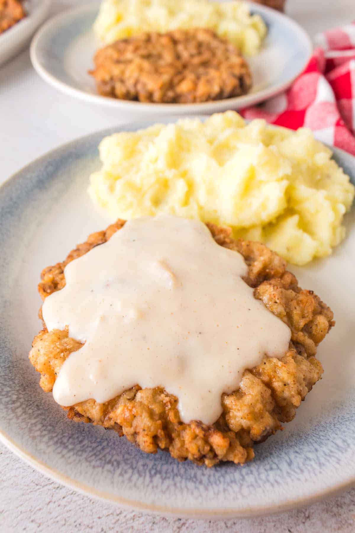 A plate of chicken fried steak, crafted from a timeless chicken fried steak recipe, is lavishly covered in white gravy and served with a generous portion of mashed potatoes. The dish is elegantly arranged on a light gray plate, with a red and white checkered cloth peeking from the background.