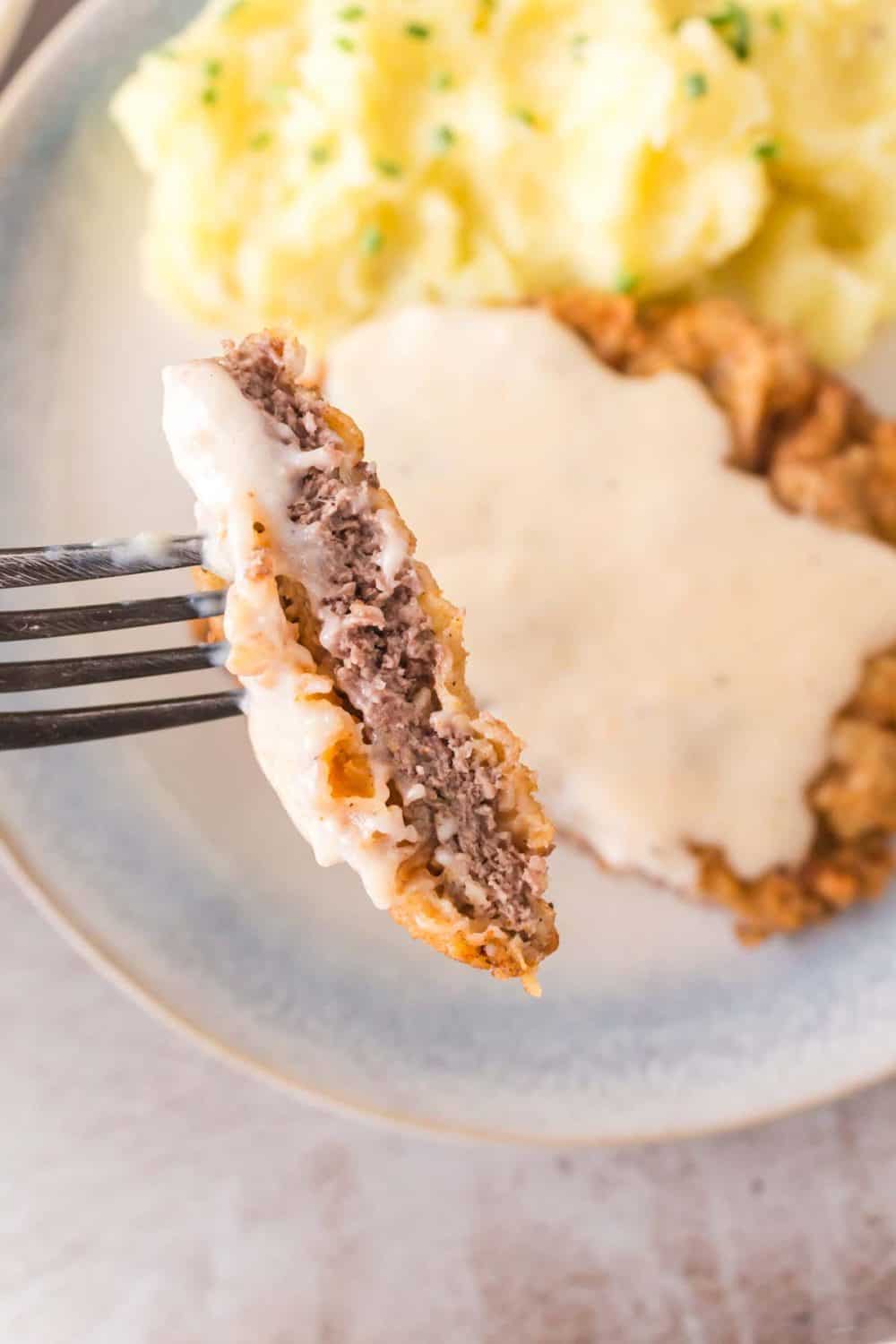A fork lifts a tantalizing piece of chicken fried steak with gravy, revealing the succulent inner meat and crispy coating. In the background, a plate showcases mashed potatoes alongside a full portion of this classic dish, drenched in rich gravy.