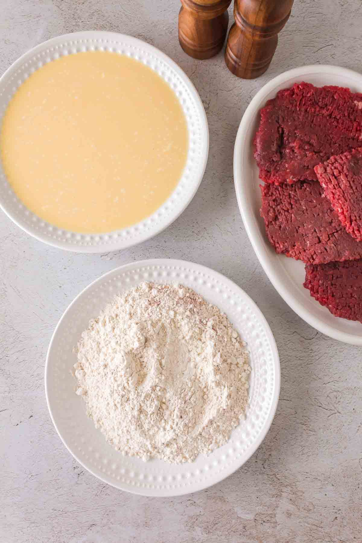 Three white plates on a light surface showcase the essentials for making chicken fried steak: one with a yellow liquid mixture, another with flour, and the third featuring pieces of raw meat. Salt and pepper grinders stand ready in the background.