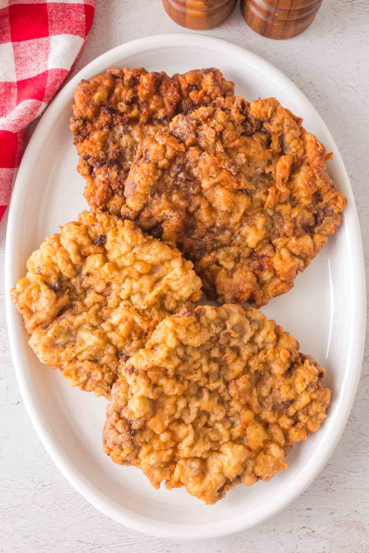 Three pieces of crispy, golden-brown chicken fried steak are stacked on a white oval plate. A red and white checkered napkin peeks from the top left corner, completing this classic comfort food presentation.