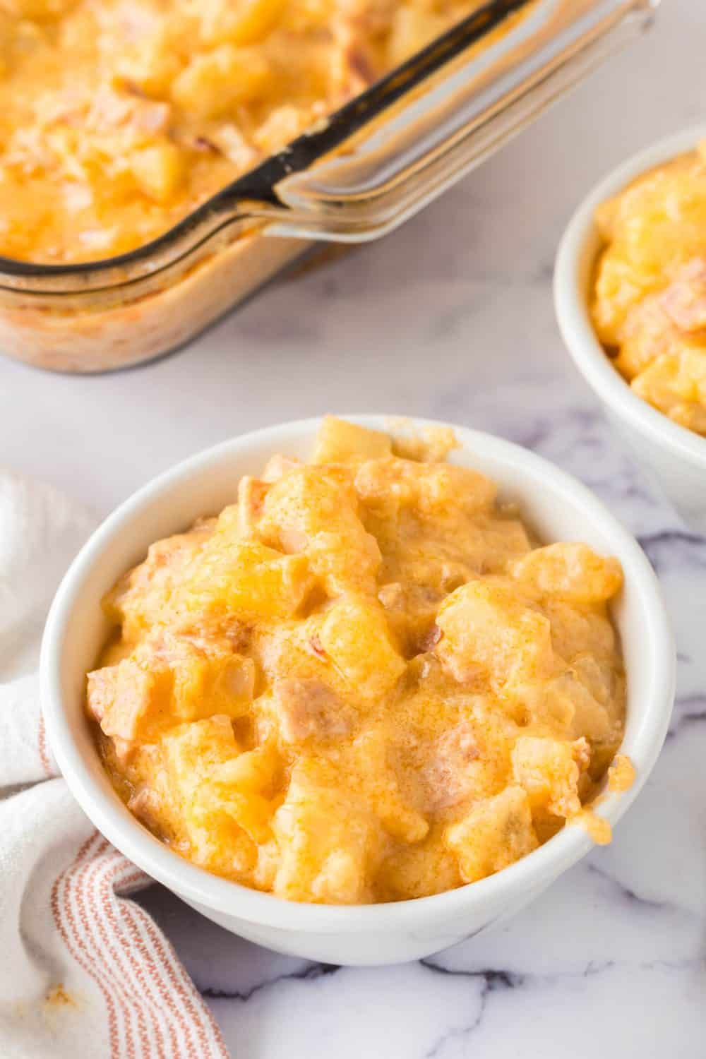 A white bowl filled with creamy, cheesy ham and potato casserole sits on a marble surface, with a baking dish and another bowl of the same casserole in the background.