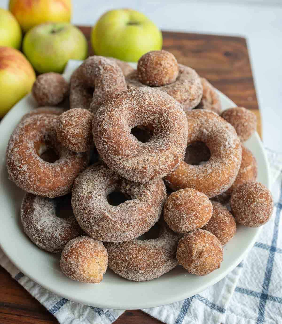A plate piled with sugar-coated apple cider donuts and donut holes sits on a wooden surface, hinting at a homemade apple cider donuts recipe, with fresh apples and a checkered cloth in the background.