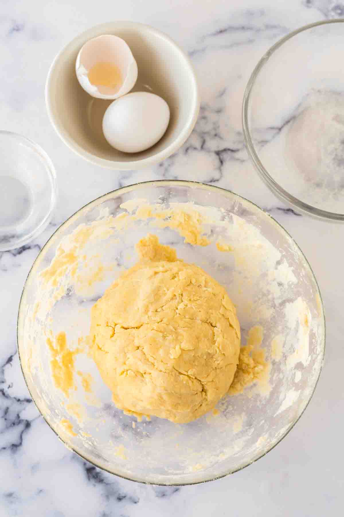 A ball of yellow empanada dough recipe sits in a glass bowl on a marble countertop, surrounded by an empty glass bowl, a small bowl with a cracked egg and eggshell, and traces of flour.