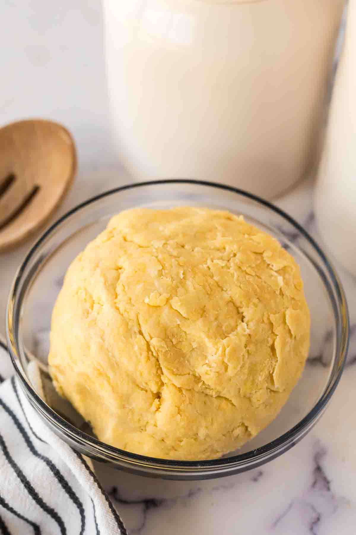 A ball of yellow empanada dough recipe sits in a clear glass bowl on a marble surface, next to a striped cloth and a wooden spoon.