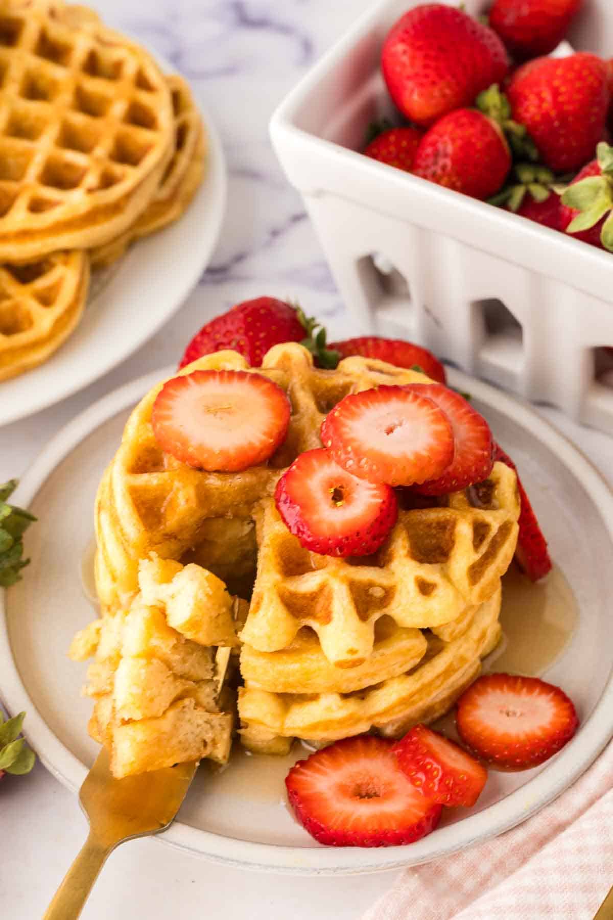 A stack of waffles from a buttermilk waffle recipe is topped with sliced strawberries and served on a white plate. A bite is cut out onto a fork, with more waffles and a basket of fresh strawberries waiting in the background.
