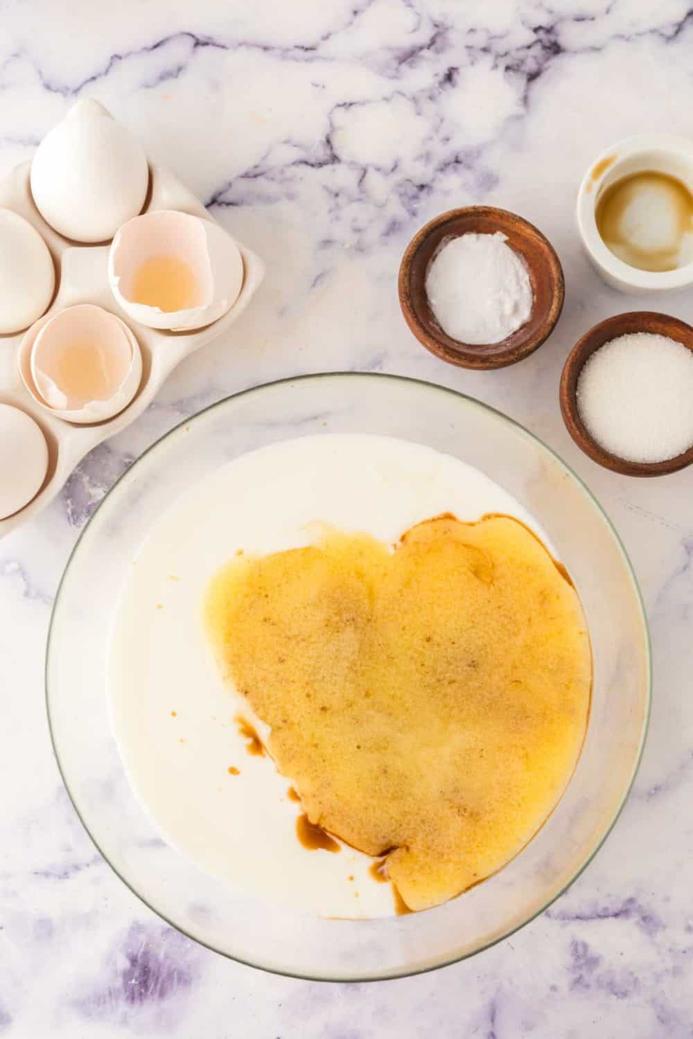 A glass bowl containing a mixture of eggs, vanilla, and milk on a marbled countertop awaits to become fluffy buttermilk waffles. Next to the bowl are cracked eggshells, a carton with eggs, and small bowls filled with baking powder and sugar.