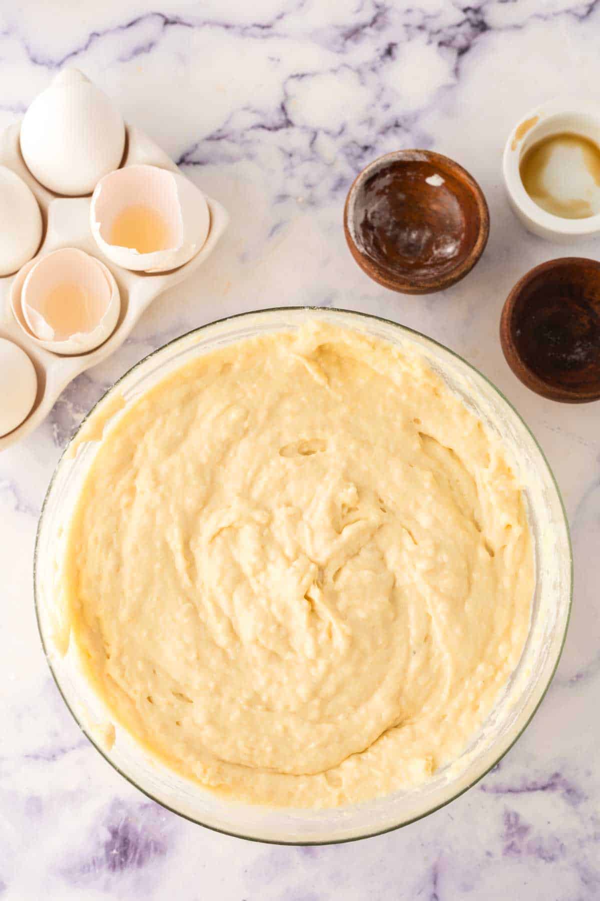 A glass bowl filled with creamy cake batter sits on a marble surface, hinting at a breakfast treat next to it: buttermilk waffles. Nearby are three small empty brown bowls, an empty white bowl, and a carton containing empty eggshells.