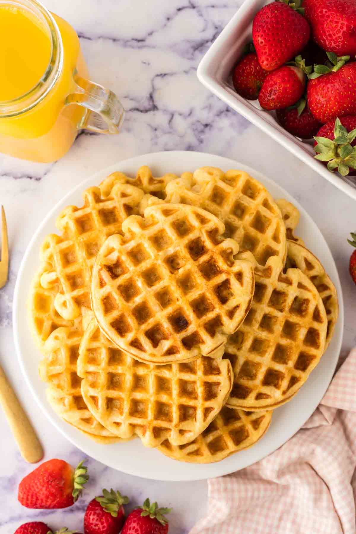 A plate of golden-brown buttermilk waffles is surrounded by a pitcher of orange juice, fresh strawberries, a knife with a wooden handle, and a pink checkered cloth on a marble surface.