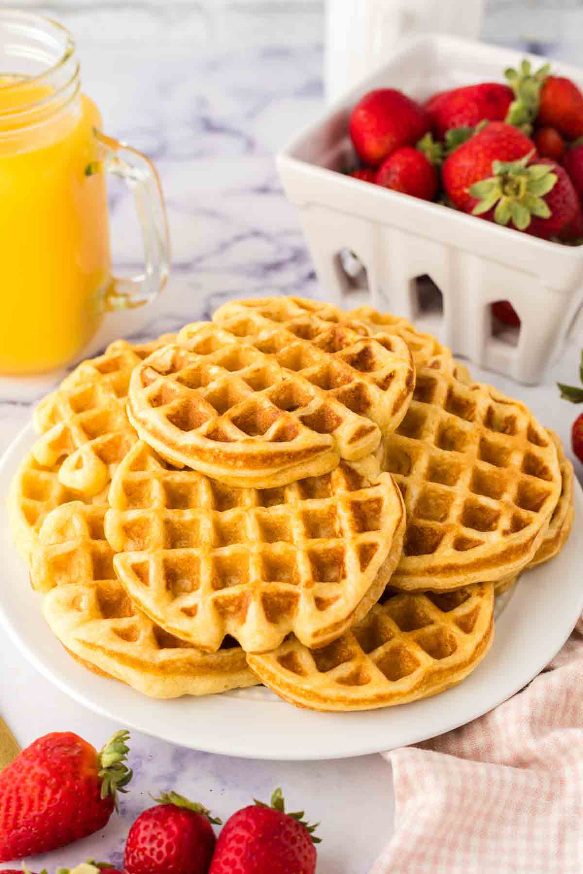 Waffles from a buttermilk waffle recipe, stacked on a white platter, surrounded by fresh strawberries. A glass mug of orange juice and a basket of strawberries grace the background, all set on a sleek marble surface.