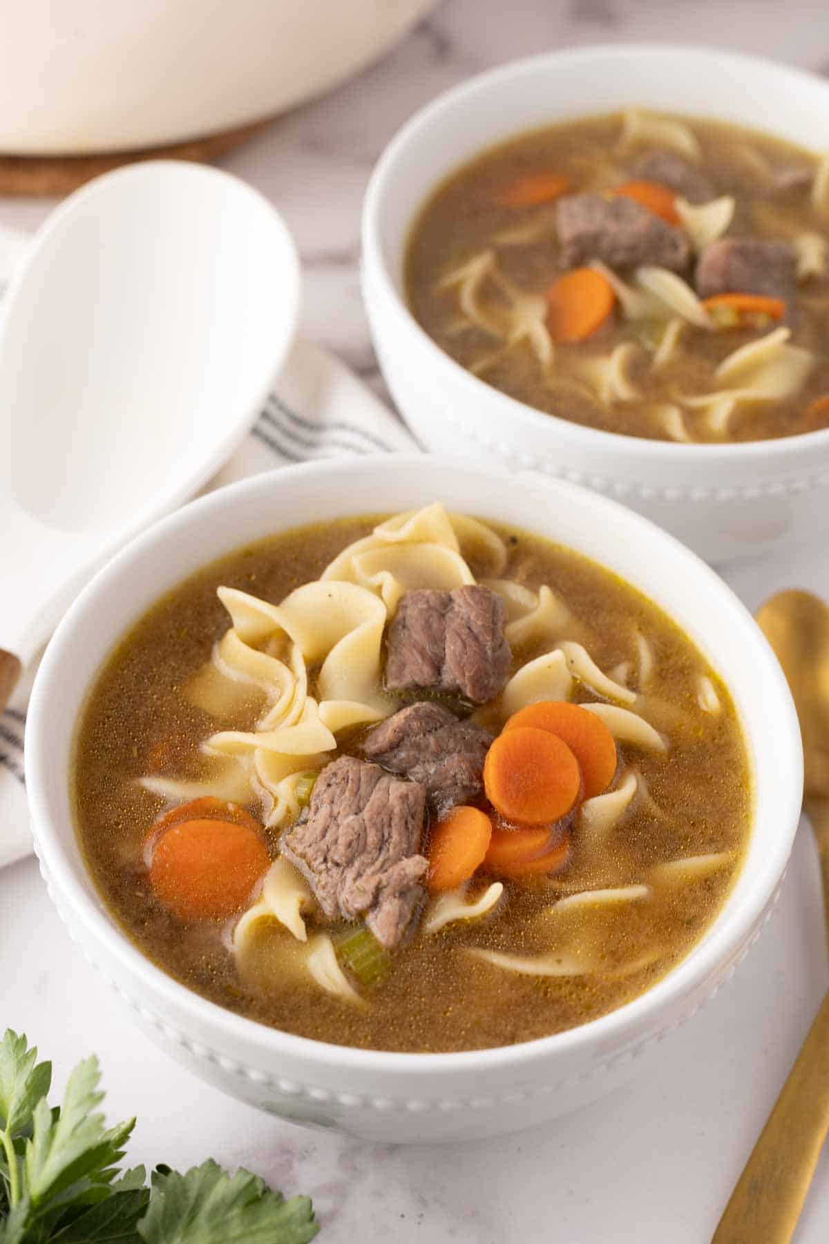 beef noodle soup recipe served in white soup bowls with a wooden spoon to the side.