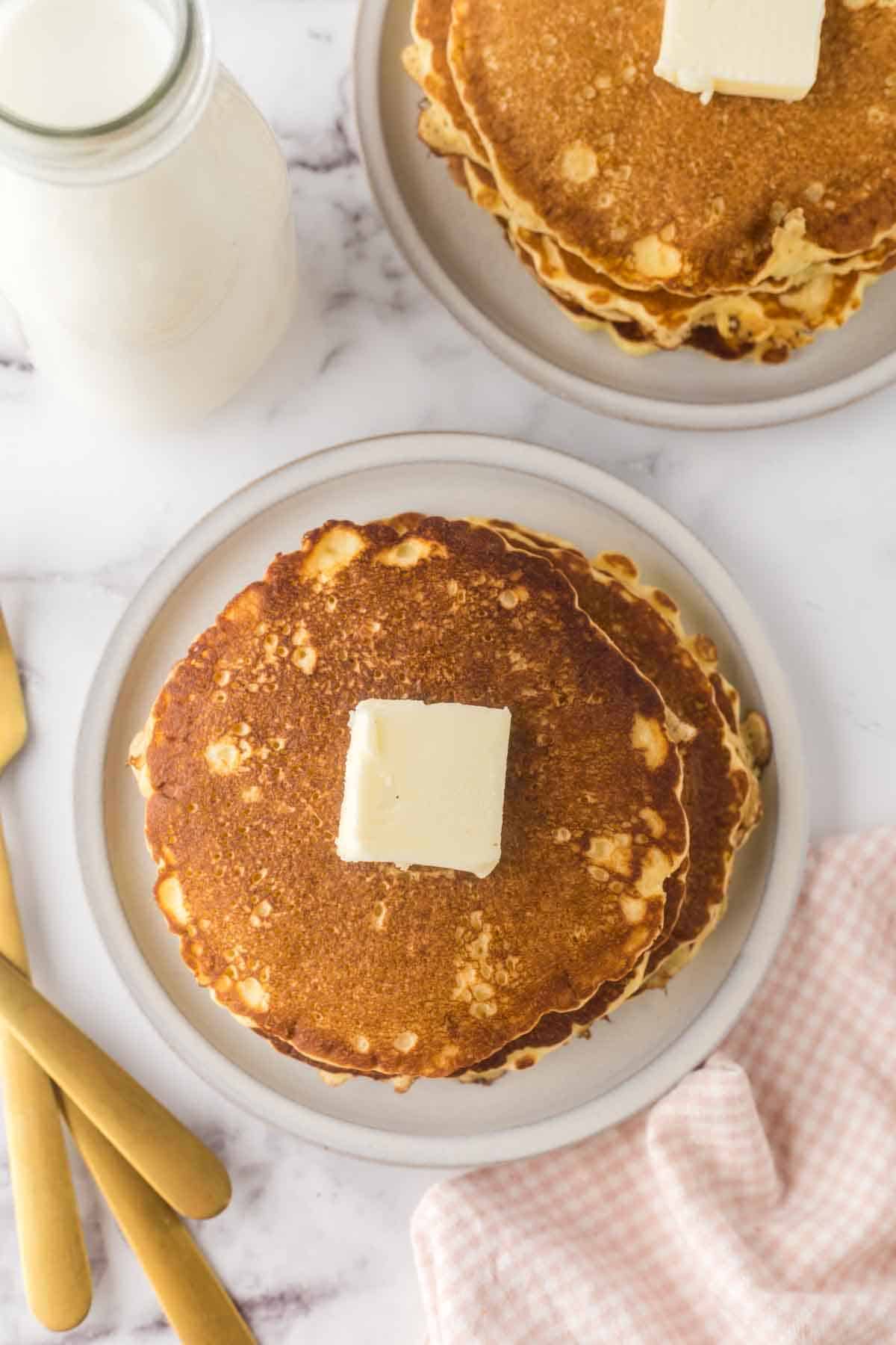 buttermilk pancakes stacked on two round plates with a pad of butter on top.