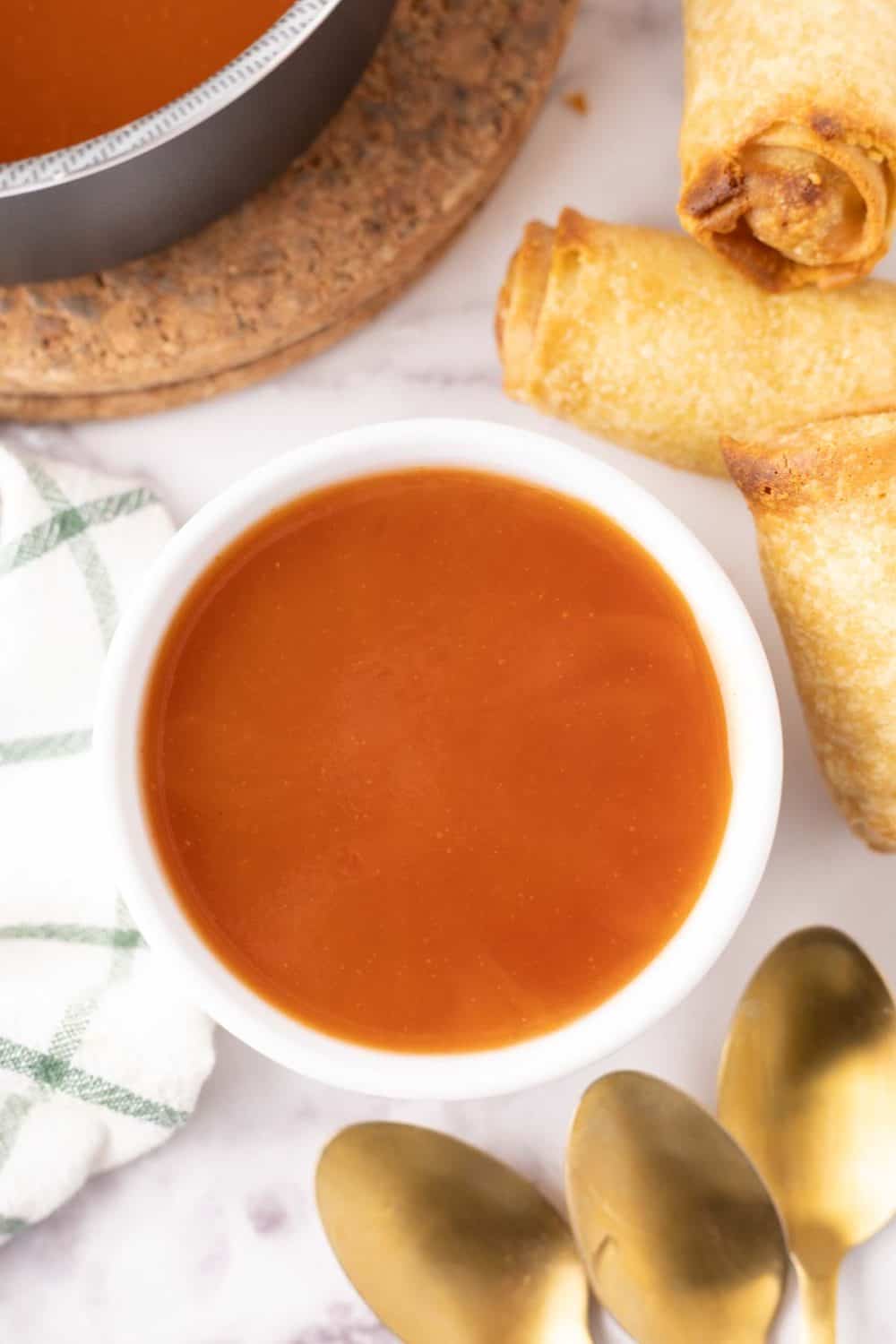 Sweet and sour sauce in a white bowl with egg rolls and gold spoons beside it.