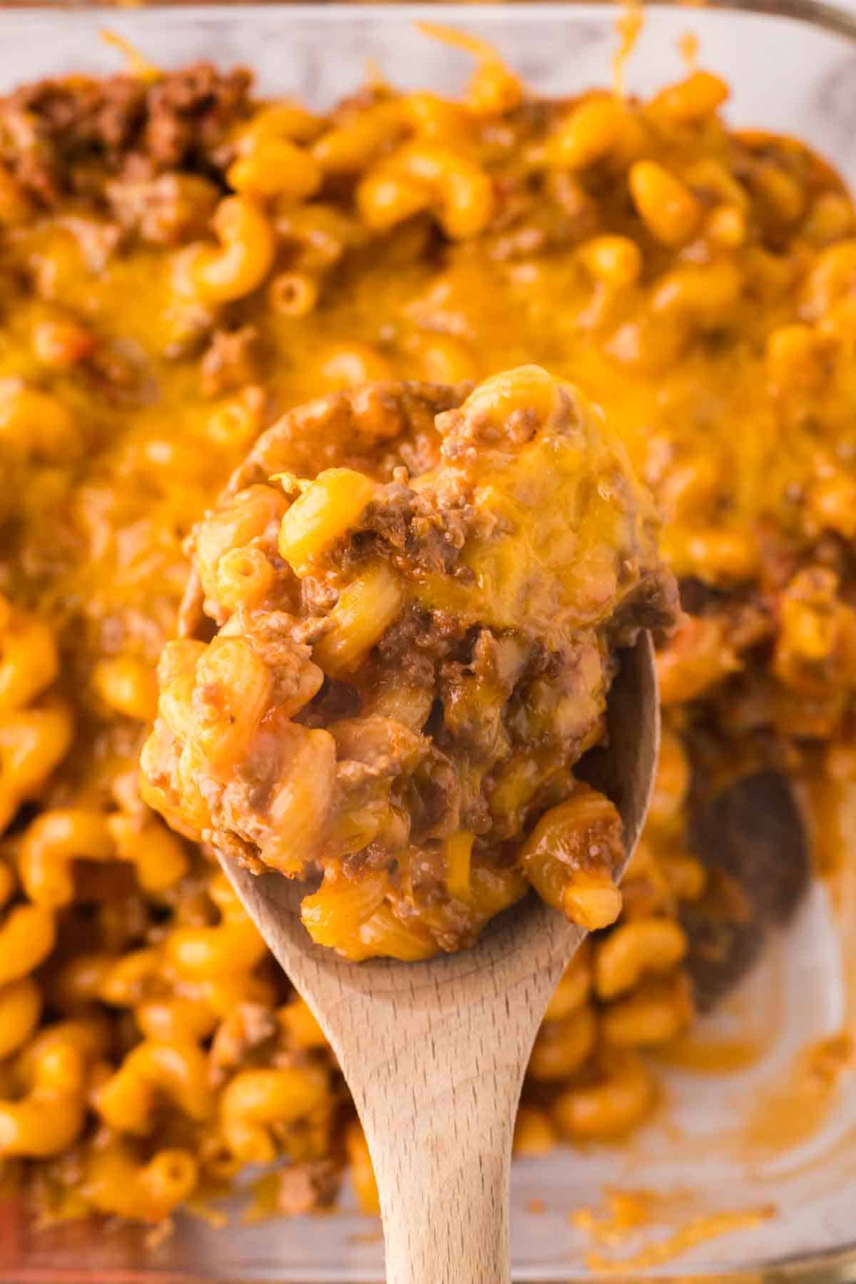 A wooden spoon holds a serving of cheesy cheeseburger casserole above a glass baking dish filled with the same hearty pasta dish.