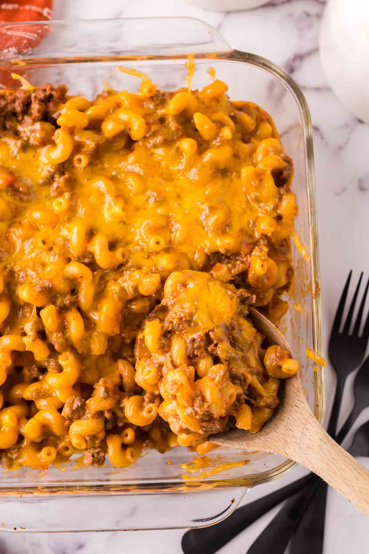 A glass baking dish filled with cheesy macaroni and ground beef, this cheeseburger casserole is topped with melted cheddar. A wooden spoon scoops out a hearty portion, while black utensils and a white napkin rest on the marble surface.