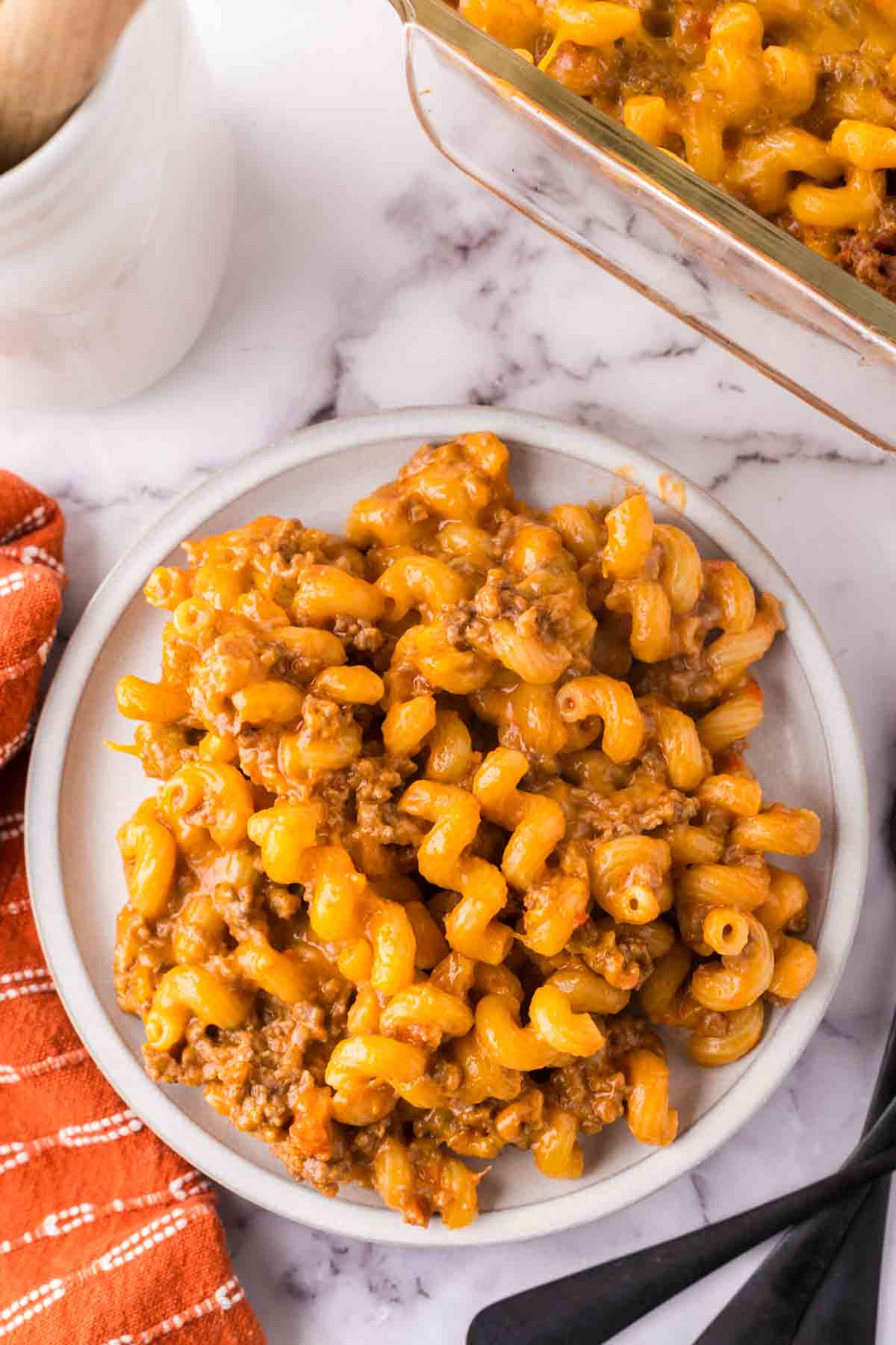 A plate of cheesy beef macaroni casserole, reminiscent of classic cheeseburger casserole, sits on a marble countertop next to an orange striped napkin, with a baking dish of the hearty meal partially visible in the corner.