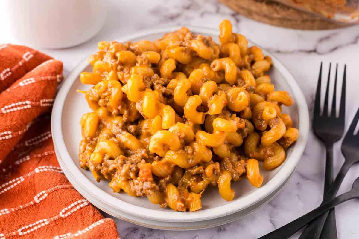 A plate of cheesy macaroni with ground beef, reminiscent of a hearty cheeseburger casserole, sits on a white surface next to two black forks and an orange napkin with white stripes.