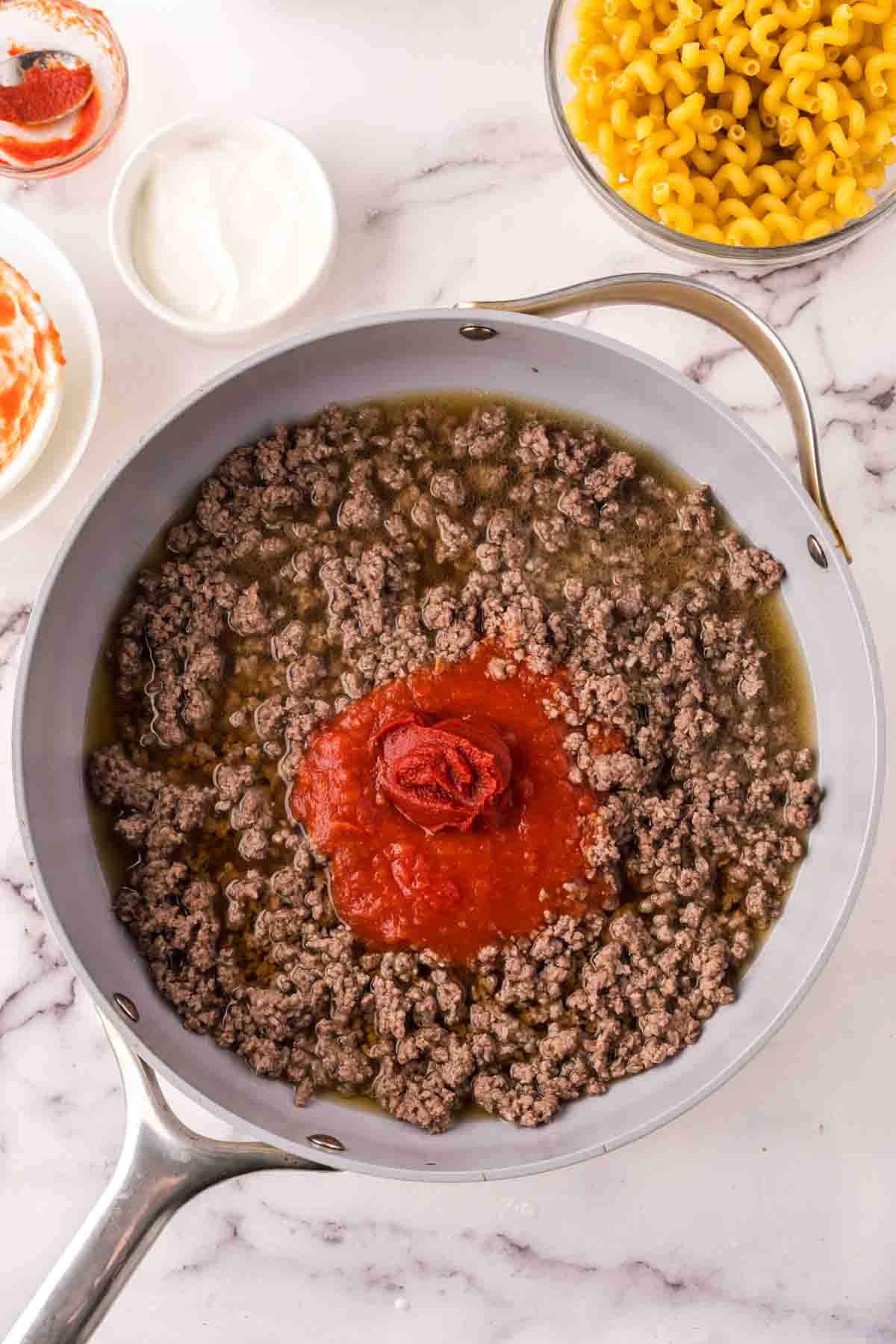 A skillet on a marble countertop contains cooked ground beef with tomato sauce and paste in the center—perfect for assembling a hearty cheeseburger casserole. Nearby are bowls of cooked macaroni, sour cream, and additional sauce.
