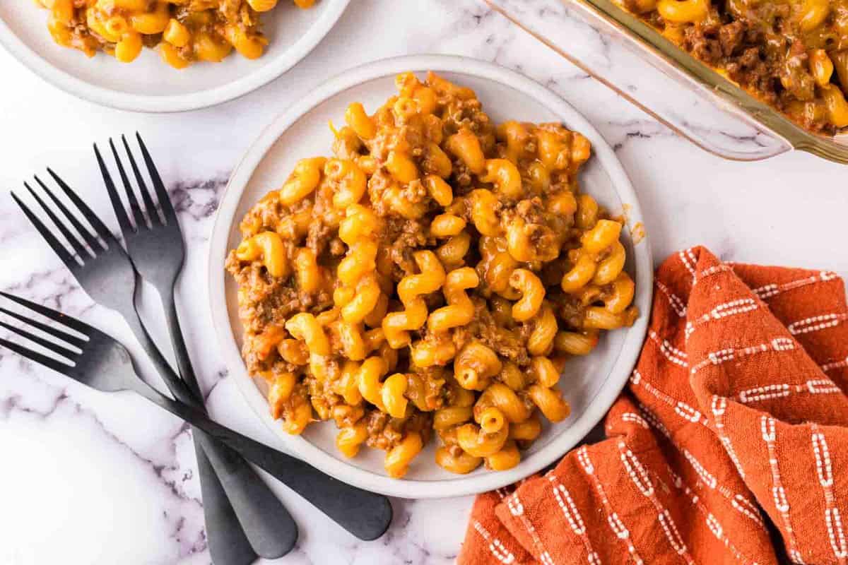 A plate of cheesy macaroni with ground beef, reminiscent of cheeseburger casserole, sits on a marble surface next to black forks and an orange checkered napkin. A casserole dish with more pasta is partially visible in the corner.