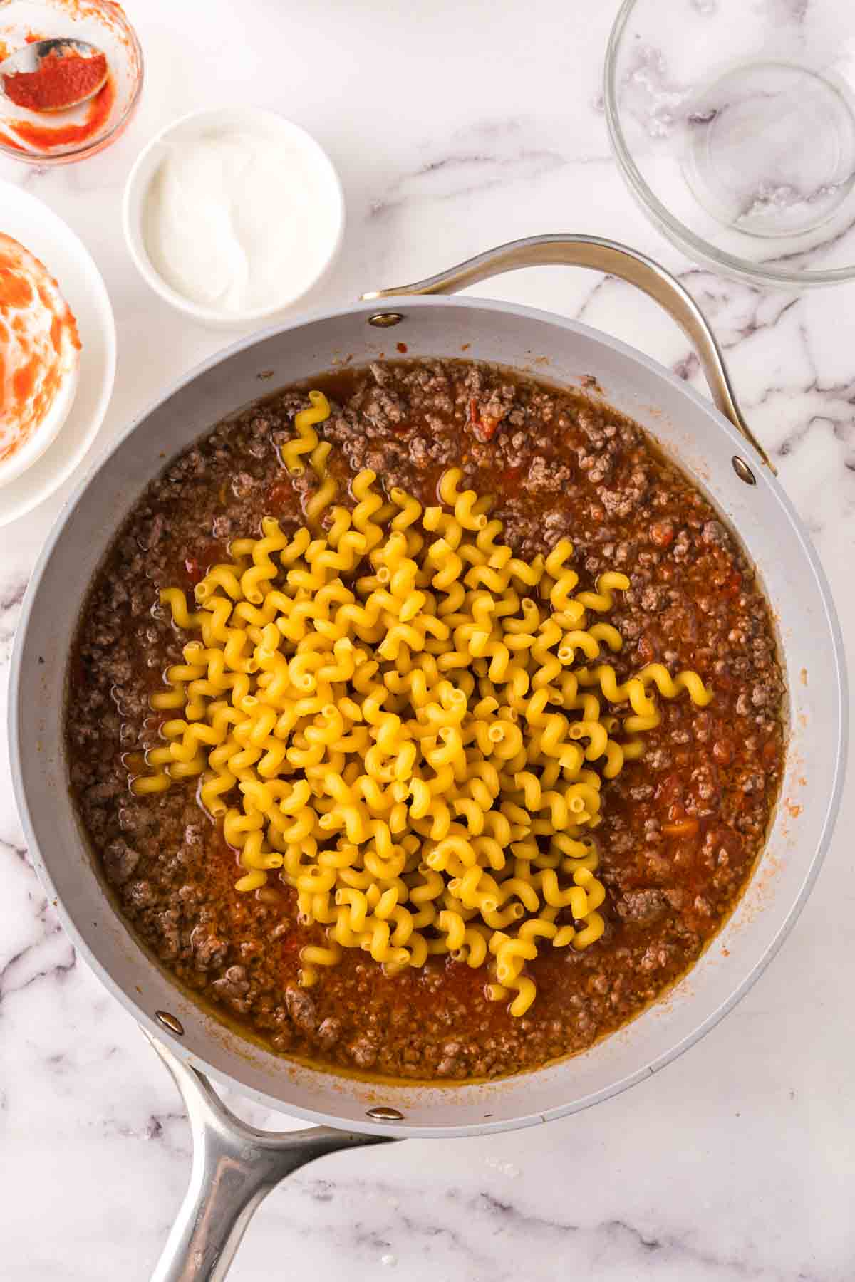 A skillet on a marble countertop filled with cooked ground beef in tomato sauce, topped with uncooked spiral pasta—perfect for a cheeseburger casserole. Small bowls with sour cream and sauce are nearby.