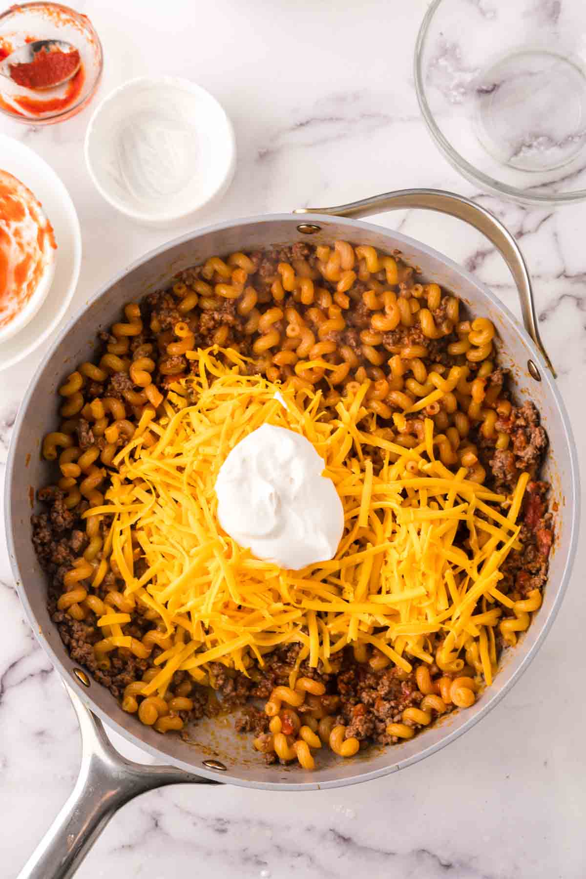 A skillet on a marble countertop filled with cheeseburger casserole—cooked macaroni, ground beef, and tomato sauce, topped with a mound of shredded cheddar cheese and a dollop of sour cream. Nearby are bowls with remnants of sauce and sour cream.