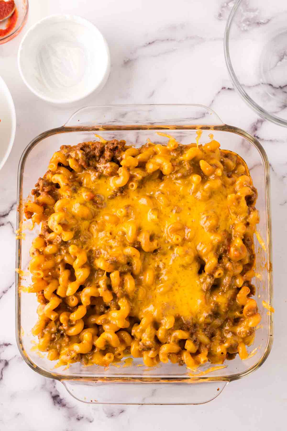 A glass baking dish filled with cheeseburger casserole—baked macaroni and ground beef, topped with melted cheddar cheese—sits on a white marble surface.