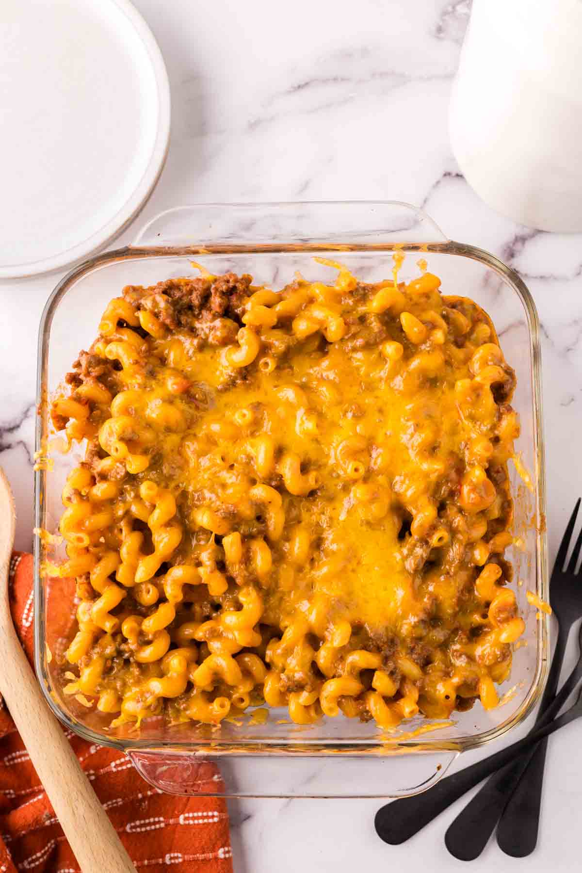 A glass baking dish filled with cheeseburger casserole—baked macaroni and cheese topped with melted cheddar and ground beef—sits next to a wooden spoon, black utensils, a red napkin, a white plate, and a glass of milk on a marble surface.