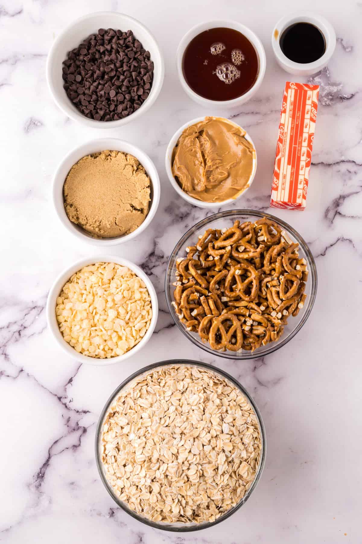 A variety of snack ingredients for healthy homemade granola bars in bowls on a marble surface, including chocolate chips, peanut butter, pretzels, oats, brown sugar, white chocolate chips, butter, vanilla extract, and syrup.