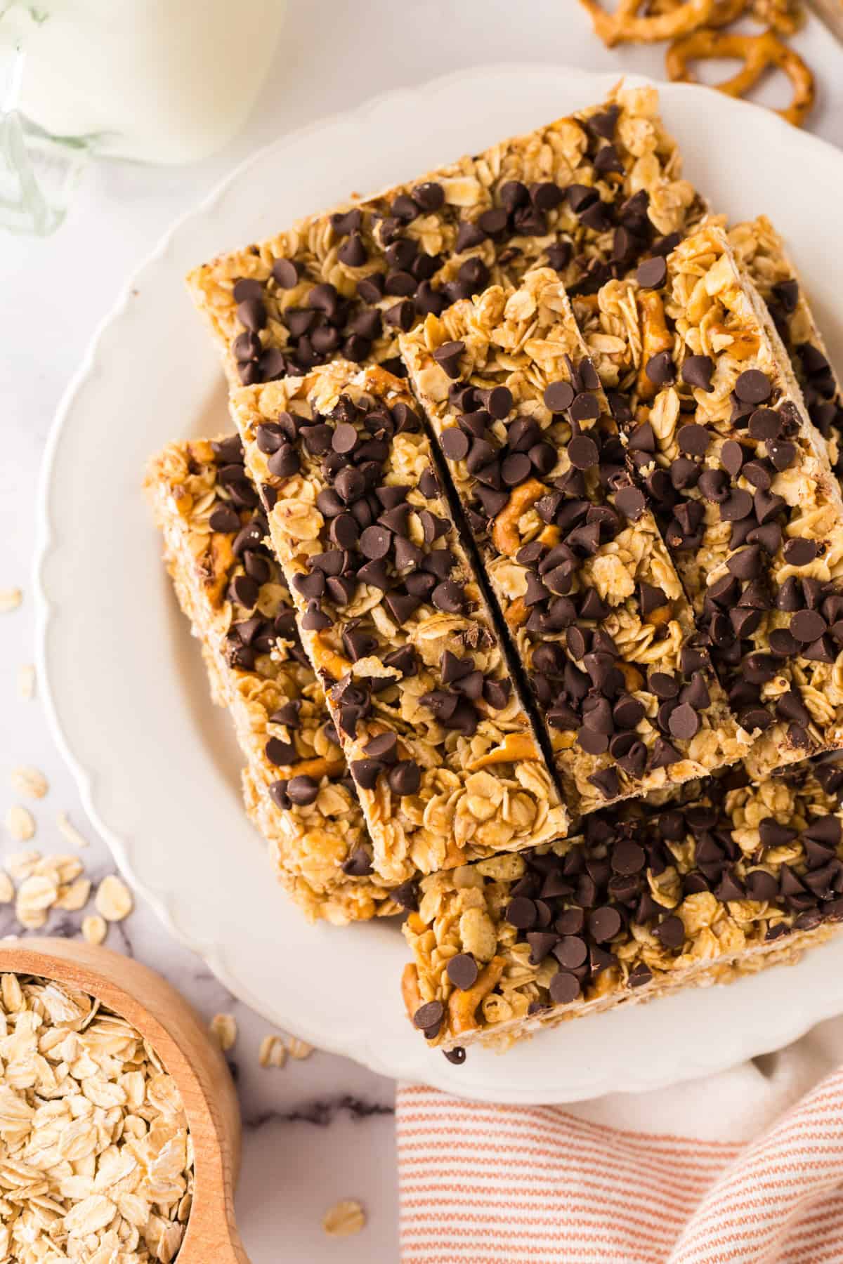 A plate stacked with several healthy homemade granola bars topped with chocolate chips. Oats and pretzel pieces are visible in the bars, with a bowl of oats and a striped cloth nearby.