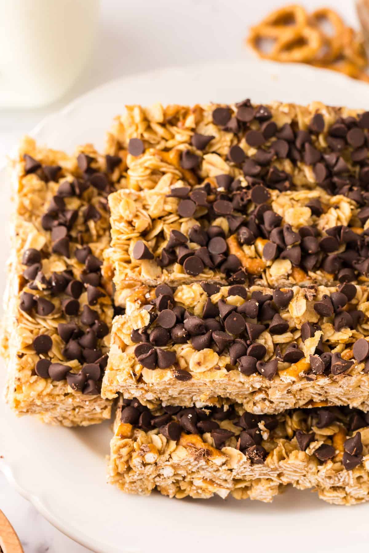 A stack of healthy homemade granola bars with oats and pretzel pieces is topped with chocolate chips on a white plate. The bars are golden brown, chunky, and perfect for a wholesome snack.