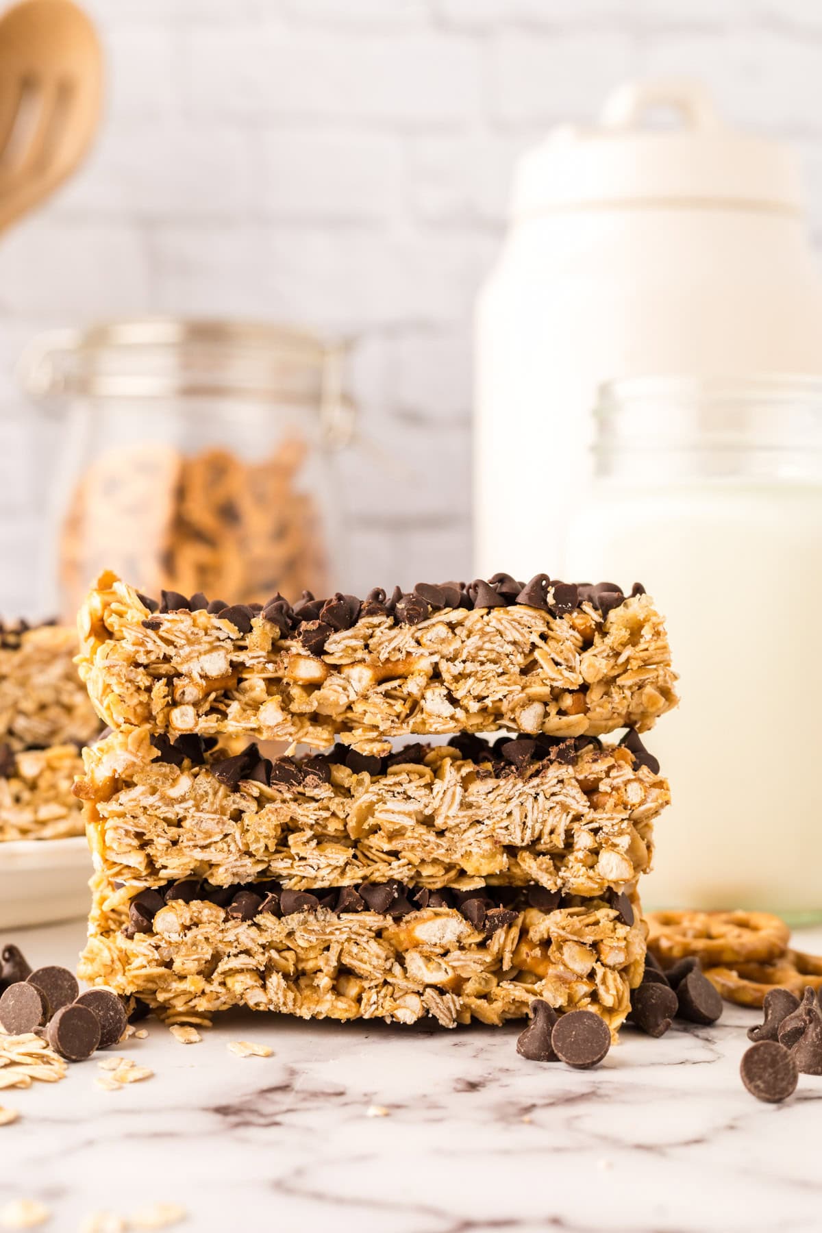 Three healthy homemade granola bars with oats and chocolate chips are stacked on a marble surface, surrounded by scattered chocolate chips. In the background, jars of milk and oats add a cozy touch, slightly out of focus.