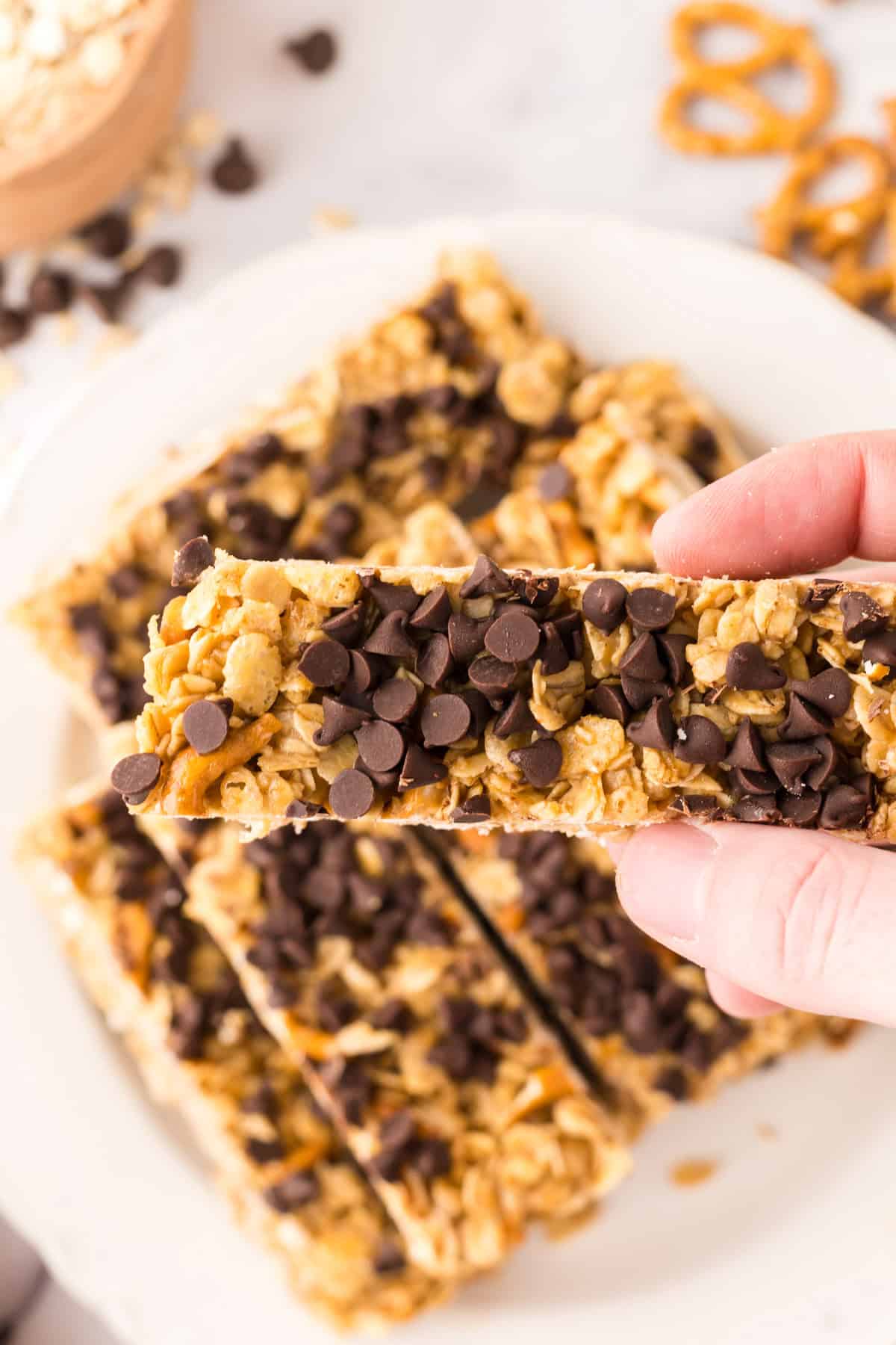A hand holds a healthy homemade granola bar topped with chocolate chips over a white plate filled with more bars. Pretzels and oats are scattered in the background.