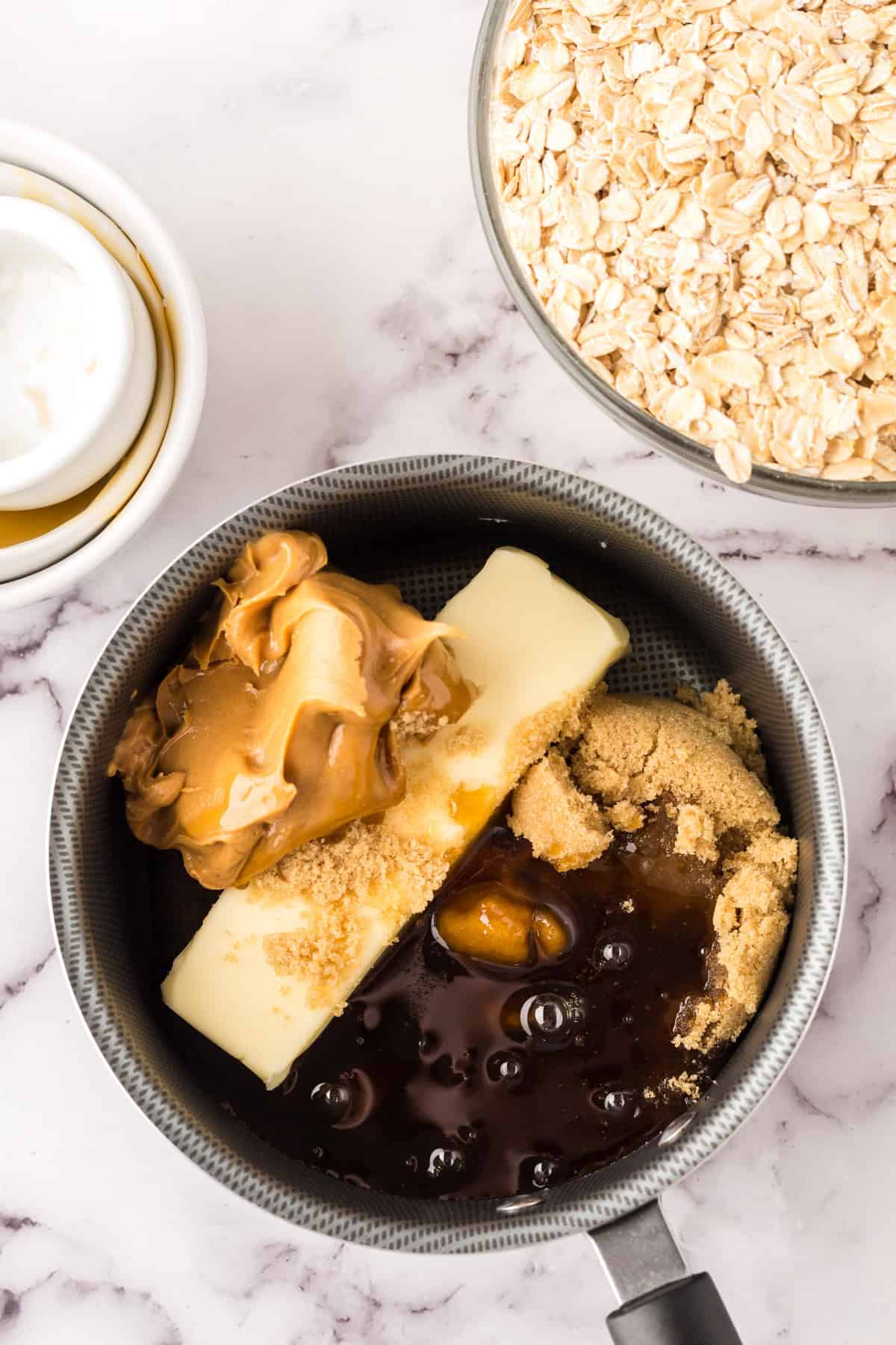 A saucepan with butter, peanut butter, brown sugar, and syrup sits on a marble surface next to a bowl of oats and a small bowl of vanilla extract—ready to make healthy homemade granola bars.