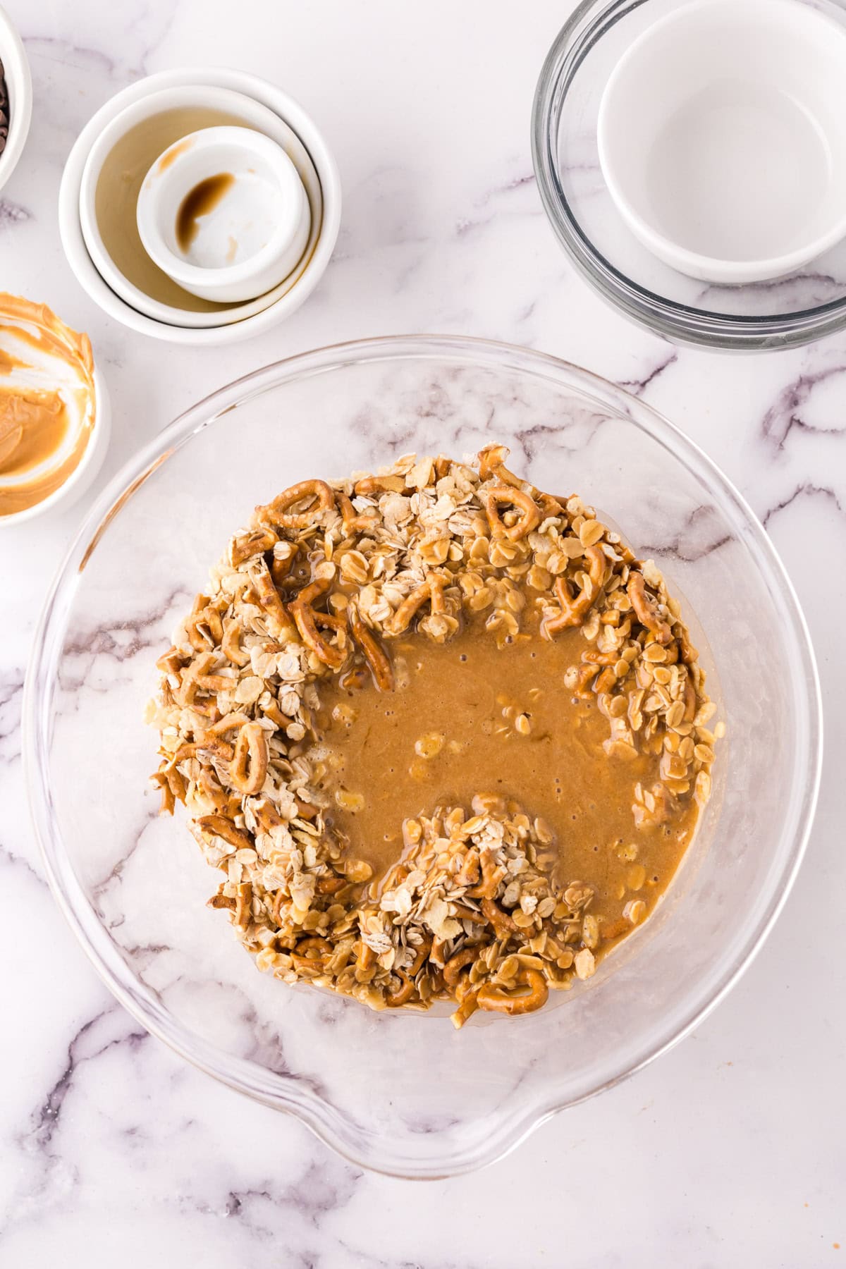 A glass mixing bowl with a mixture of oats, crushed pretzels, and peanut butter for healthy homemade granola bars sits on a marble countertop. Surrounding it are small bowls with remnants of ingredients and empty white dishes.