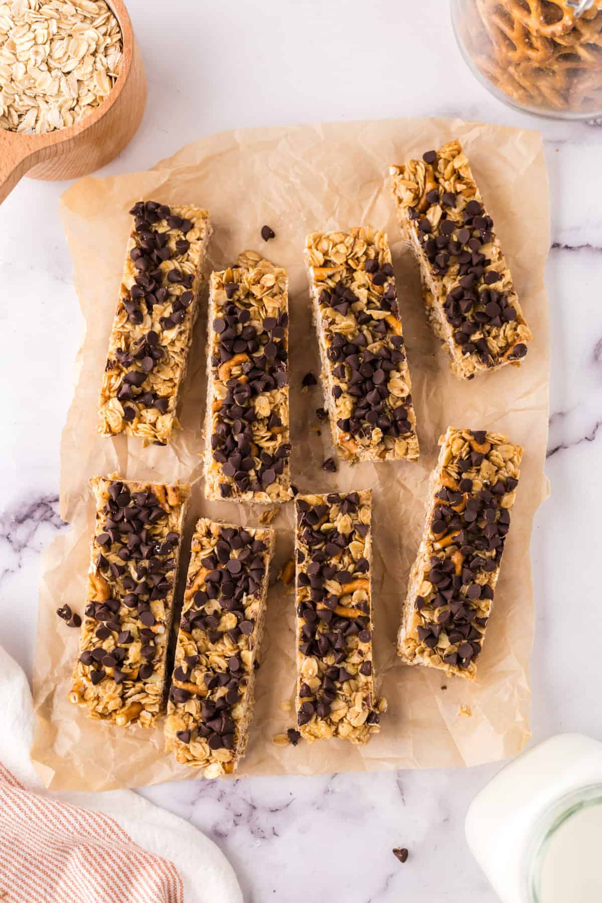 Eight healthy homemade granola bars topped with chocolate chips and oats are arranged on parchment paper, with a bowl of oats and a jar of milk nearby on a marble surface.