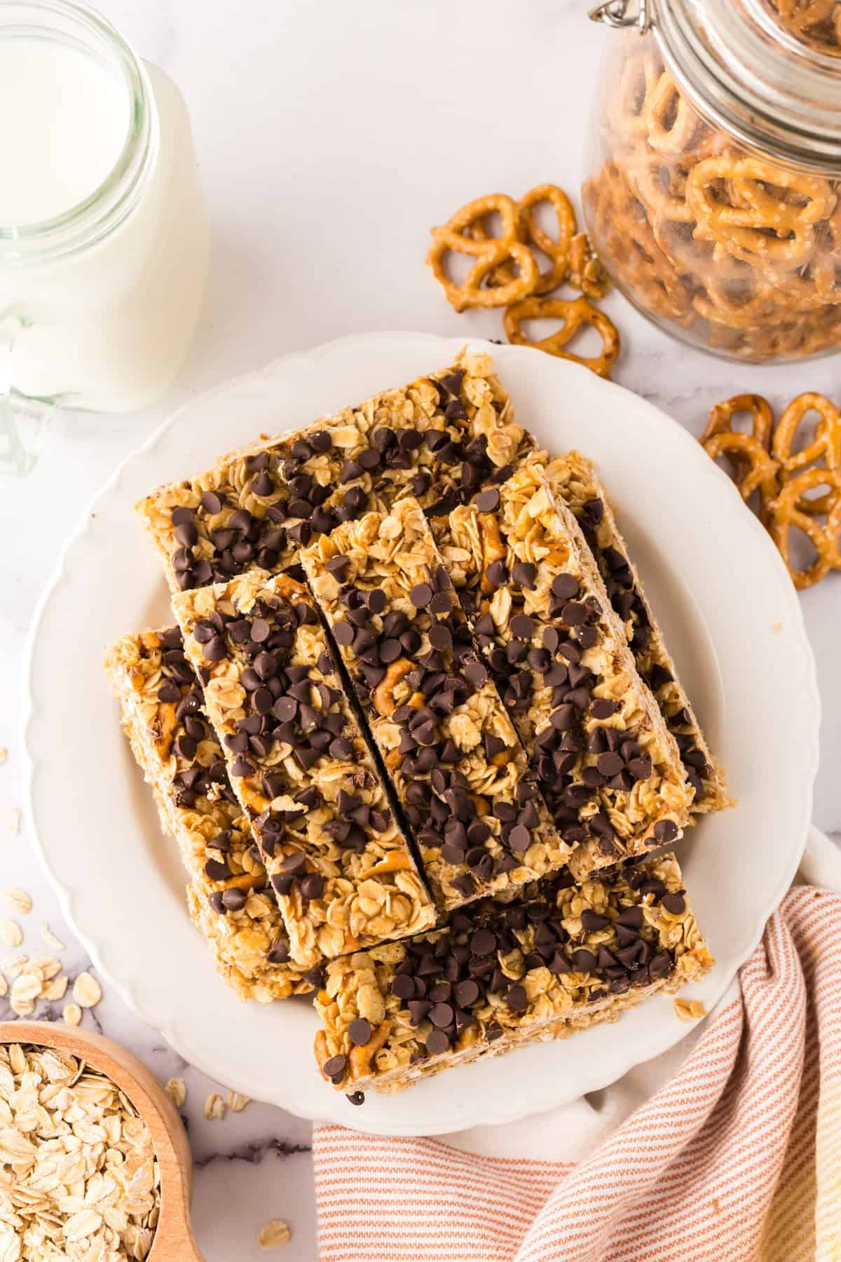 A plate of healthy homemade granola bars topped with chocolate chips sits next to a glass of milk, a jar of pretzels, and a bowl of oats on a marble surface.