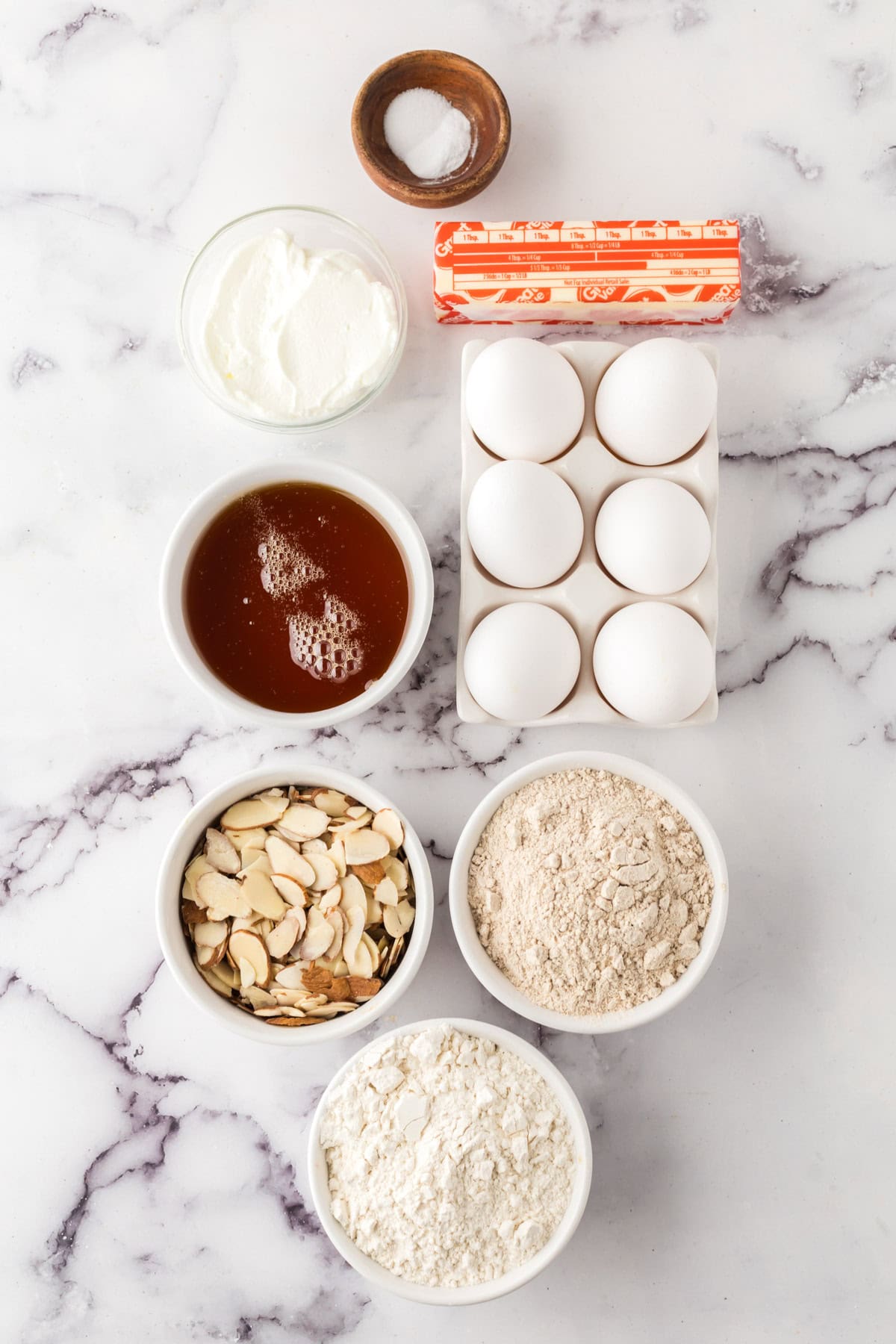A marble countertop displays bowls of sliced almonds, honey, flour, whole wheat flour, salt, butter, eggs, and sour cream—all neatly arranged in rows for a delicious honey cake recipe.