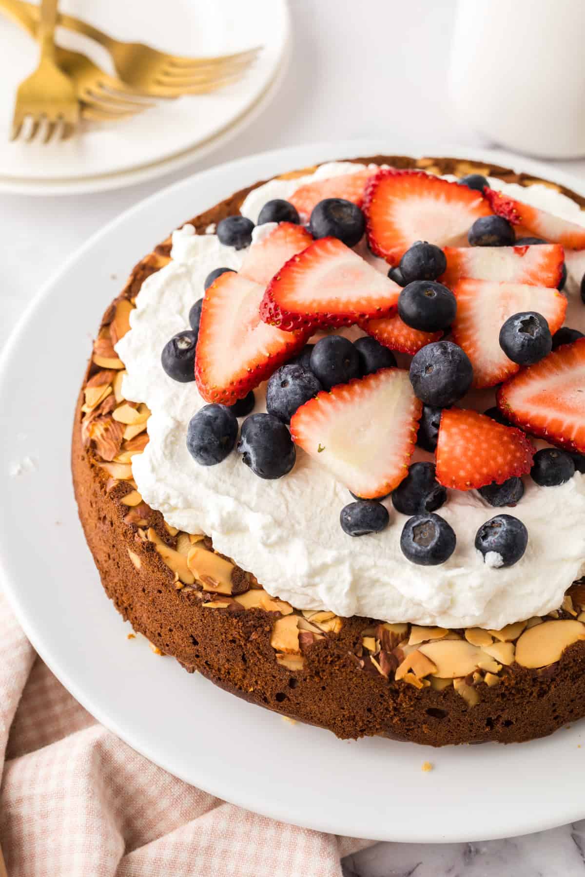 A round honey cake recipe topped with whipped cream, sliced strawberries, and blueberries sits on a white plate. A stack of plates with gold forks and a glass of milk are in the background.