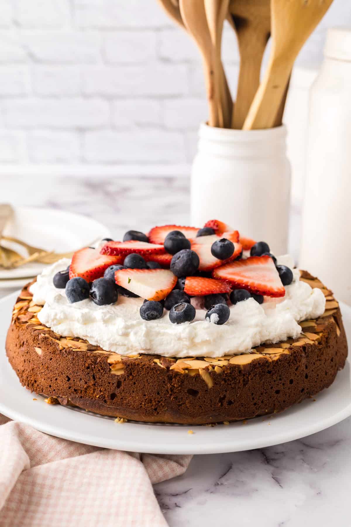 A round honey cake recipe topped with whipped cream, sliced strawberries, and blueberries sits on a white plate. Wooden utensils in a white jar and a milk bottle are in the background on a marble countertop.
