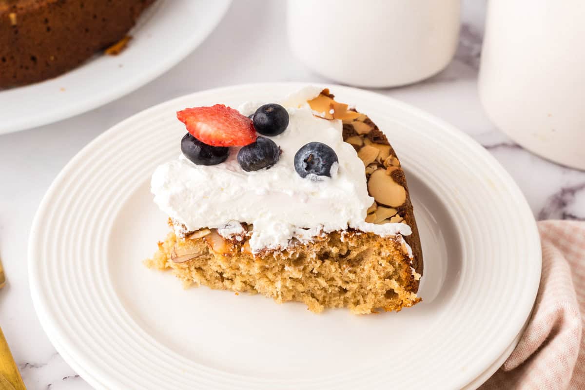 A slice of honey cake inspired by a honey cake recipe is topped with whipped cream, blueberries, and a sliced strawberry on a white plate. The cake has a light, fluffy texture and almond slices on top.