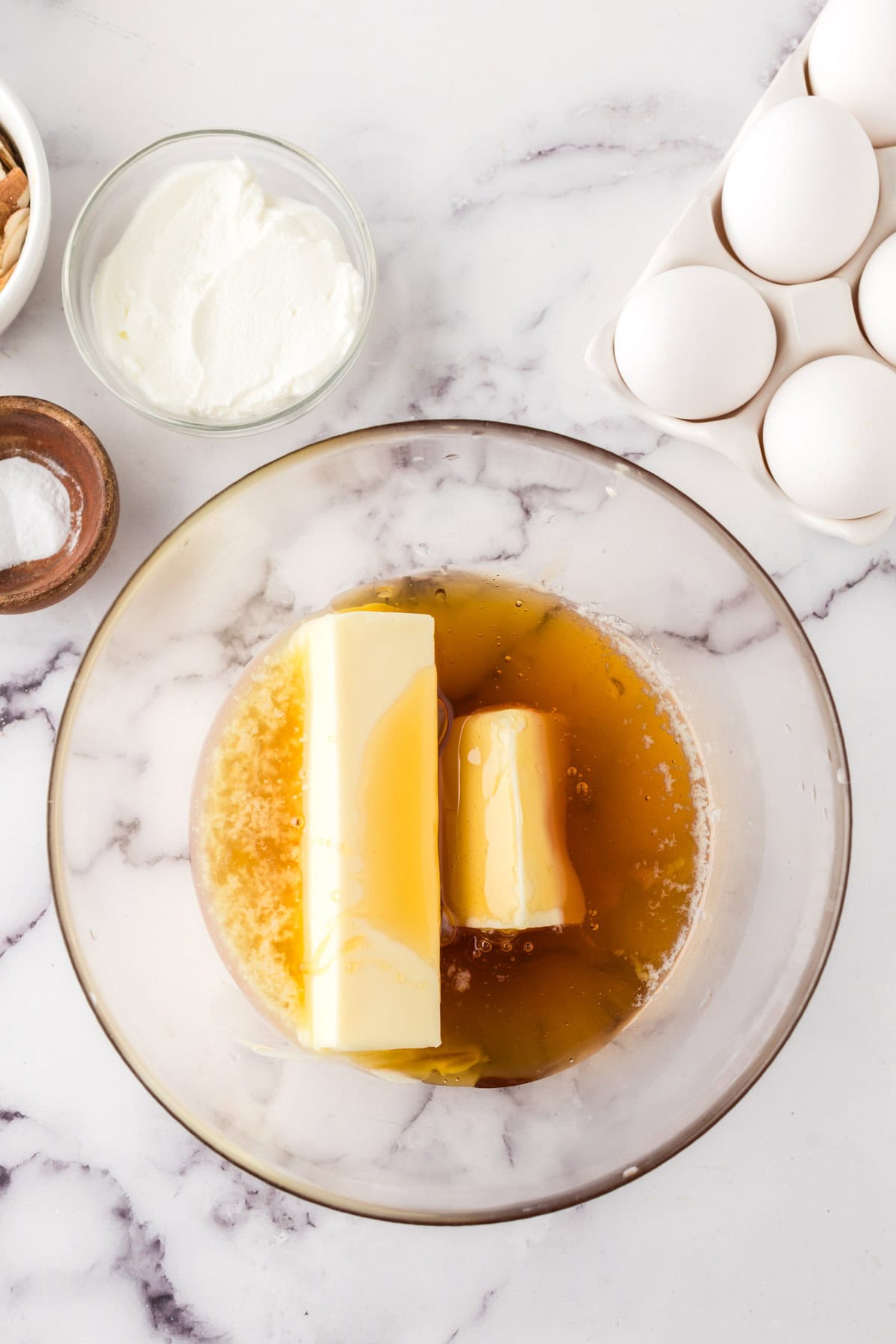 A glass bowl contains two sticks of butter and honey, perfect for a honey cake recipe. Nearby on the marble counter are eggs in a carton, a small bowl of sour cream, almonds, and salt.