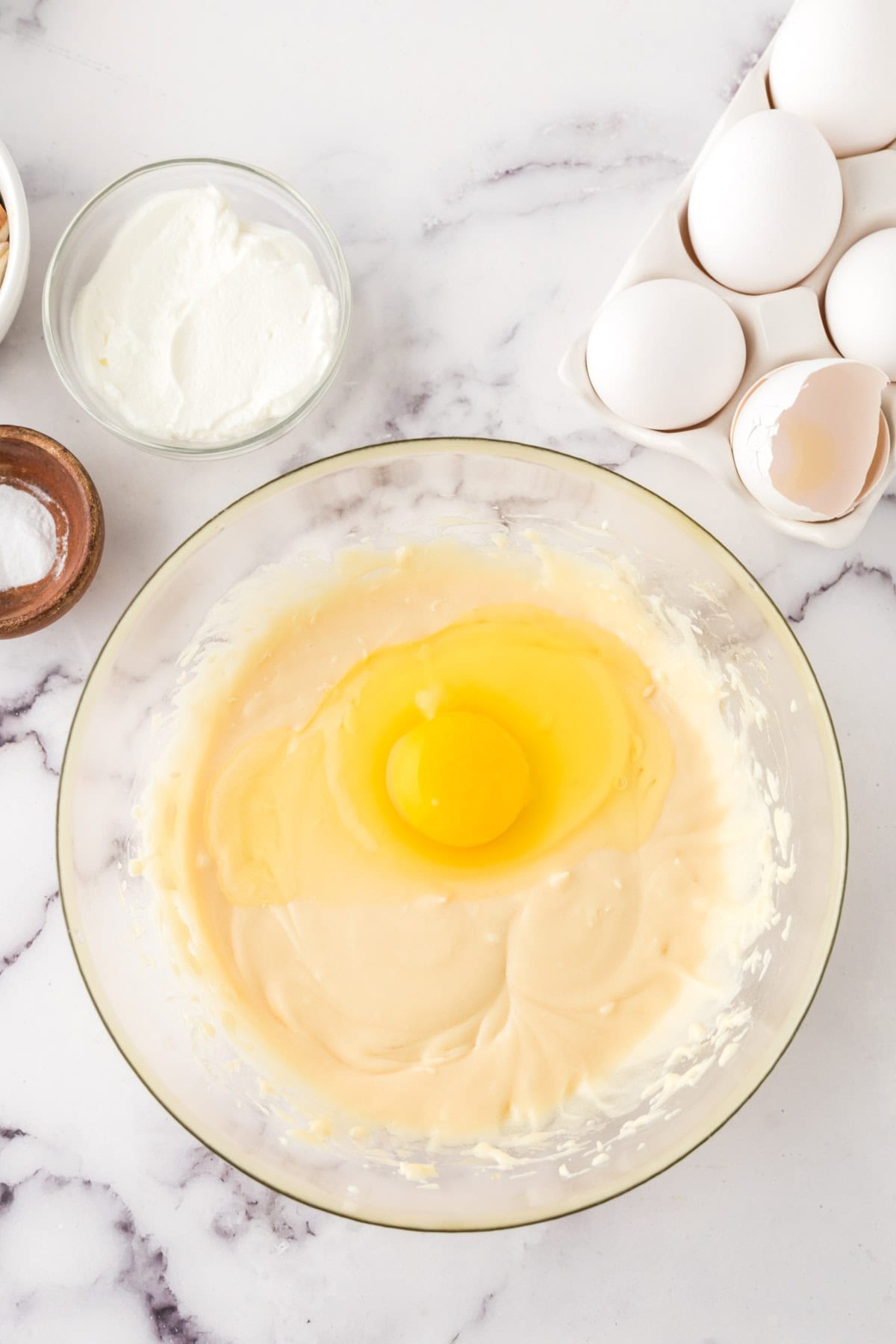 A glass bowl with creamy batter and a raw egg on top sits on a marble surface, surrounded by ingredients for a honey cake recipe—a bowl of white cream, a small dish of salt, and a carton of eggs, one cracked open.