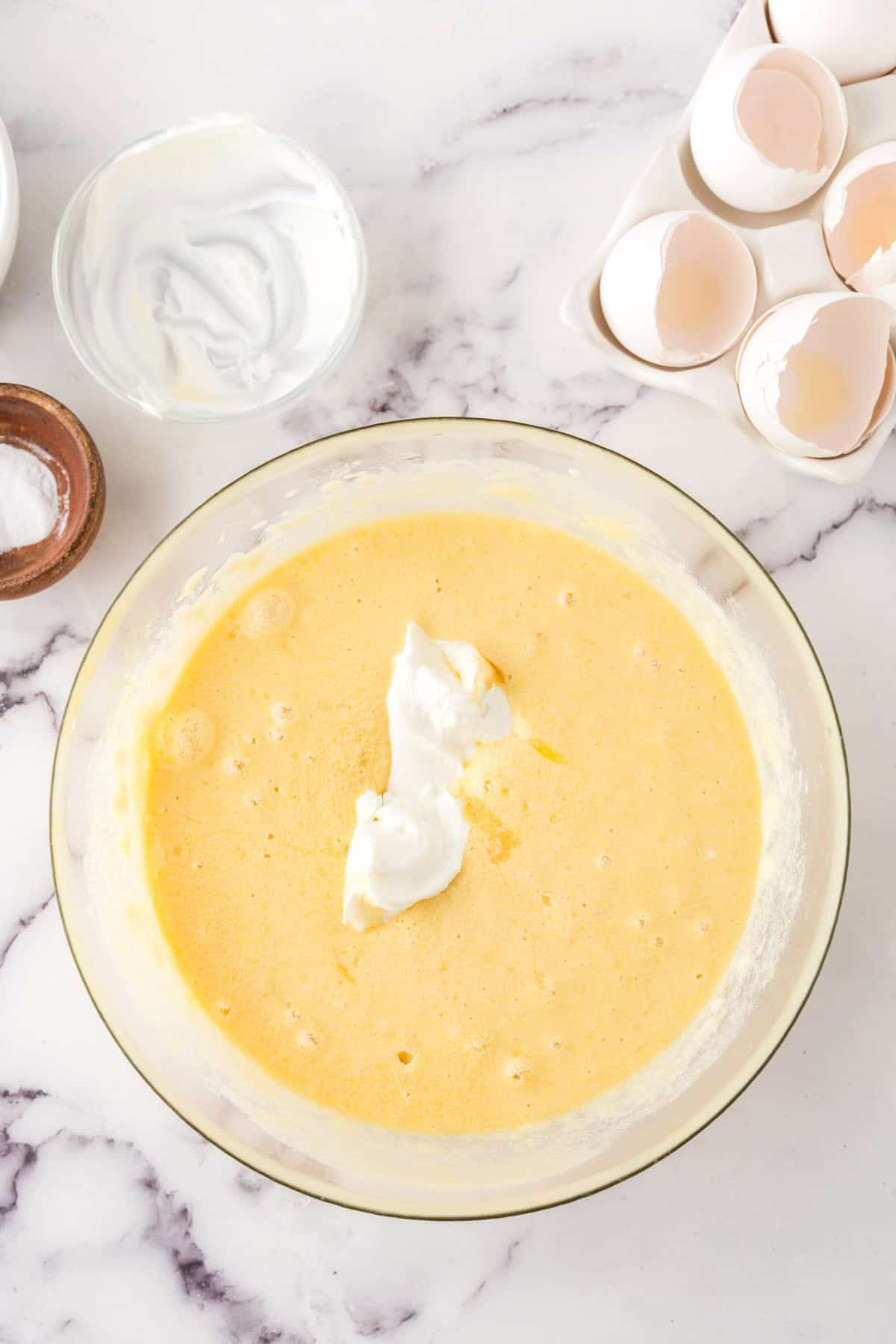 A glass mixing bowl filled with yellow cake batter for a honey cake recipe, topped with a dollop of sour cream, surrounded by empty eggshells, a small bowl of sour cream, and a small bowl of salt on a marble countertop.